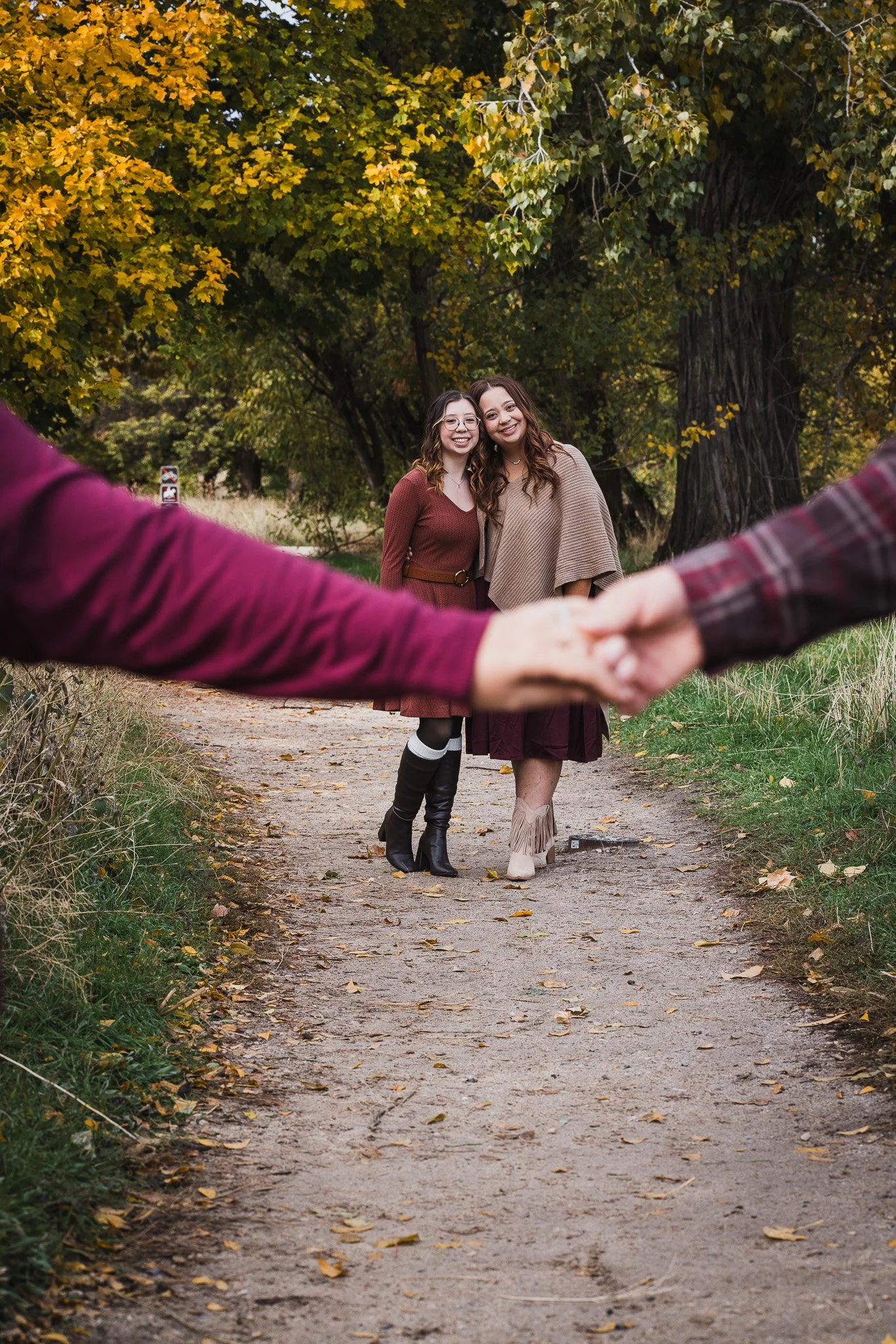 Two women holding hands and smiling, standing on a dirt path in a park with autumn foliage.
