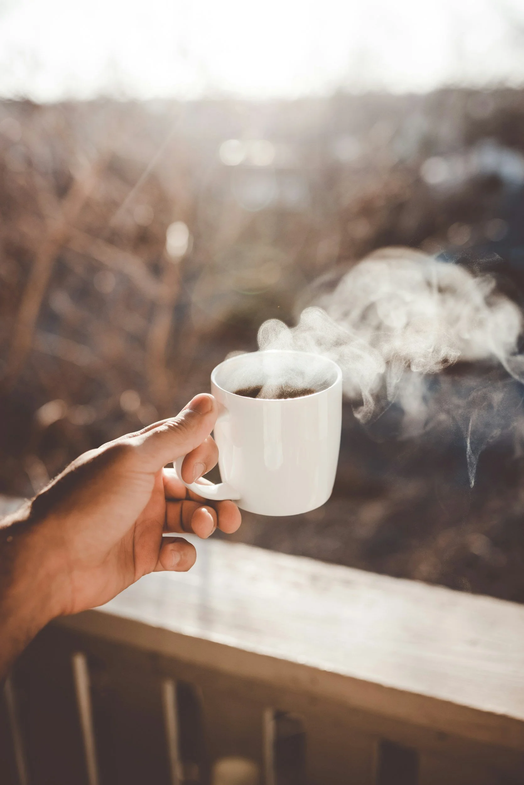 A person's hand holding a white mug filled with a hot beverage, with steam rising from it, near a window with sunlight.