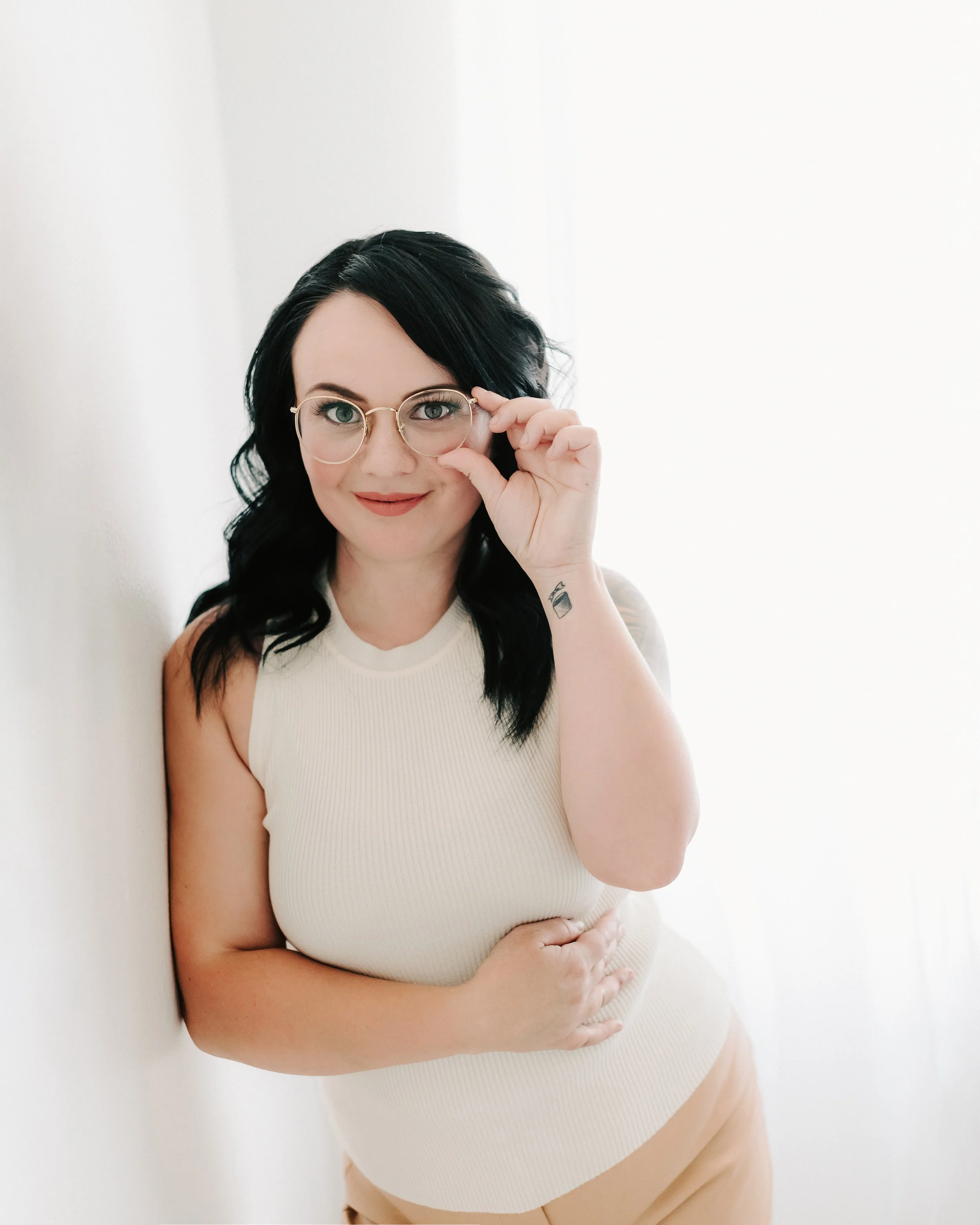 A woman with dark hair and glasses, smiling and holding her glasses with her right hand, standing against a plain white background.