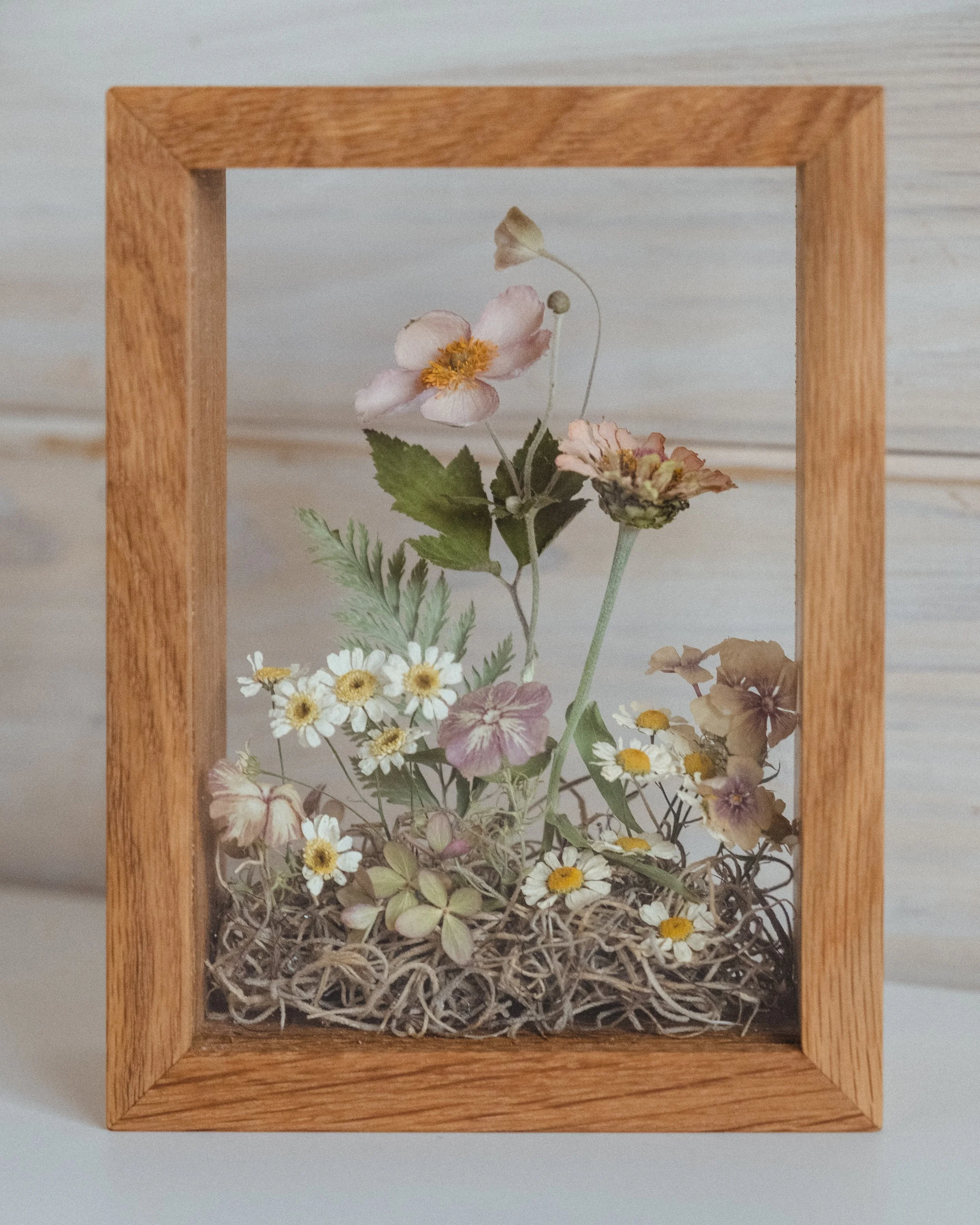 A collection of dried flowers inside a glass case with a wooden frame, set against a light background.