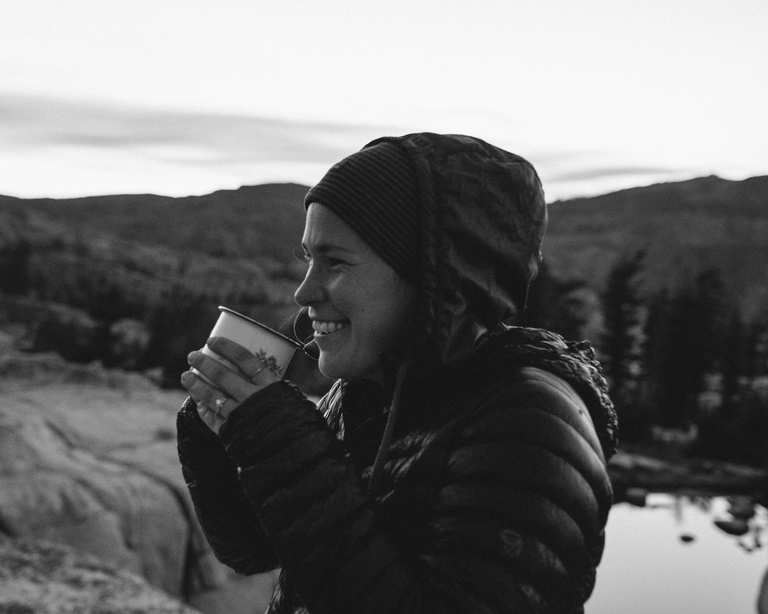 A woman with a beanie and puffy jacket is smiling and holding a cup, standing outdoors near a lake with a mountainous landscape in the background during sunset.