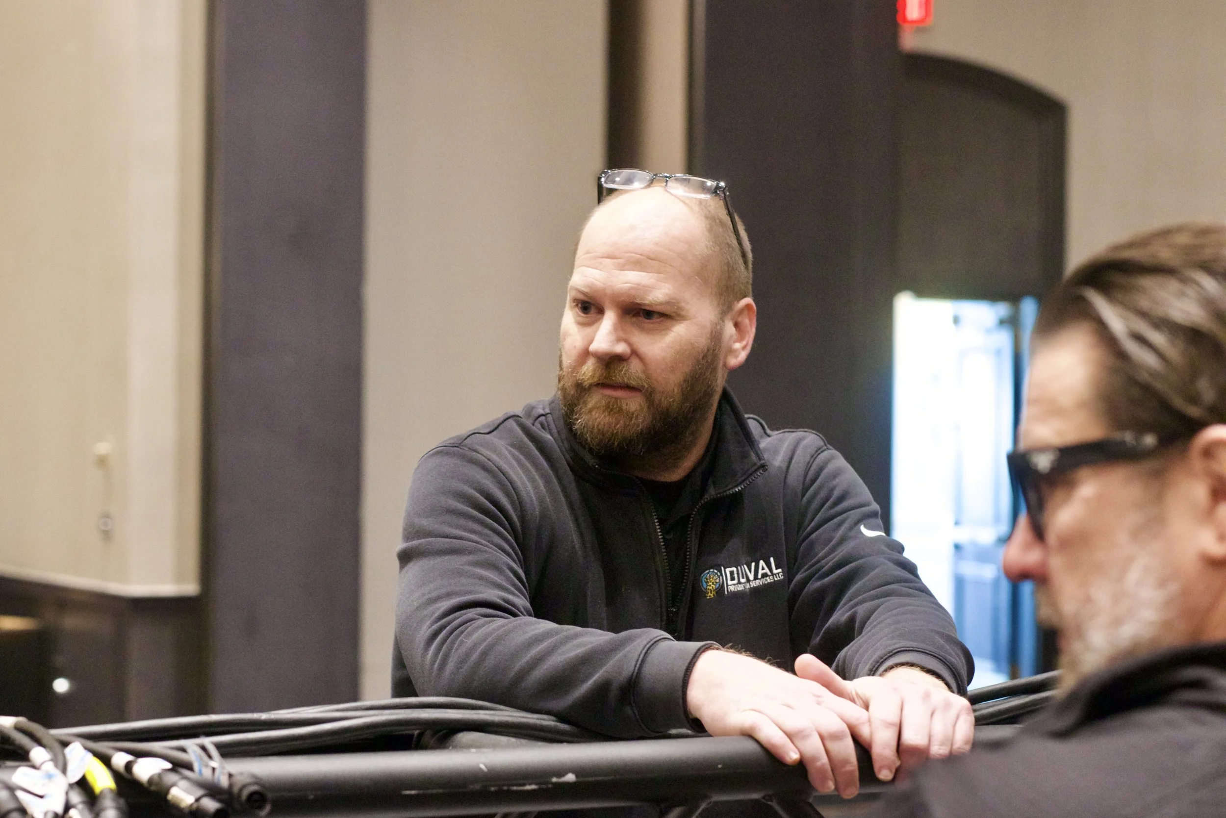 A man with a beard and glasses on his forehead, wearing a black jacket, sitting at a table with wires and electronic components, looking to his right.
