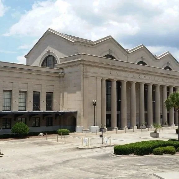 A large, historic building with tall columns and arched windows, likely a government or civic building, with landscaping and a clear sky in the background.