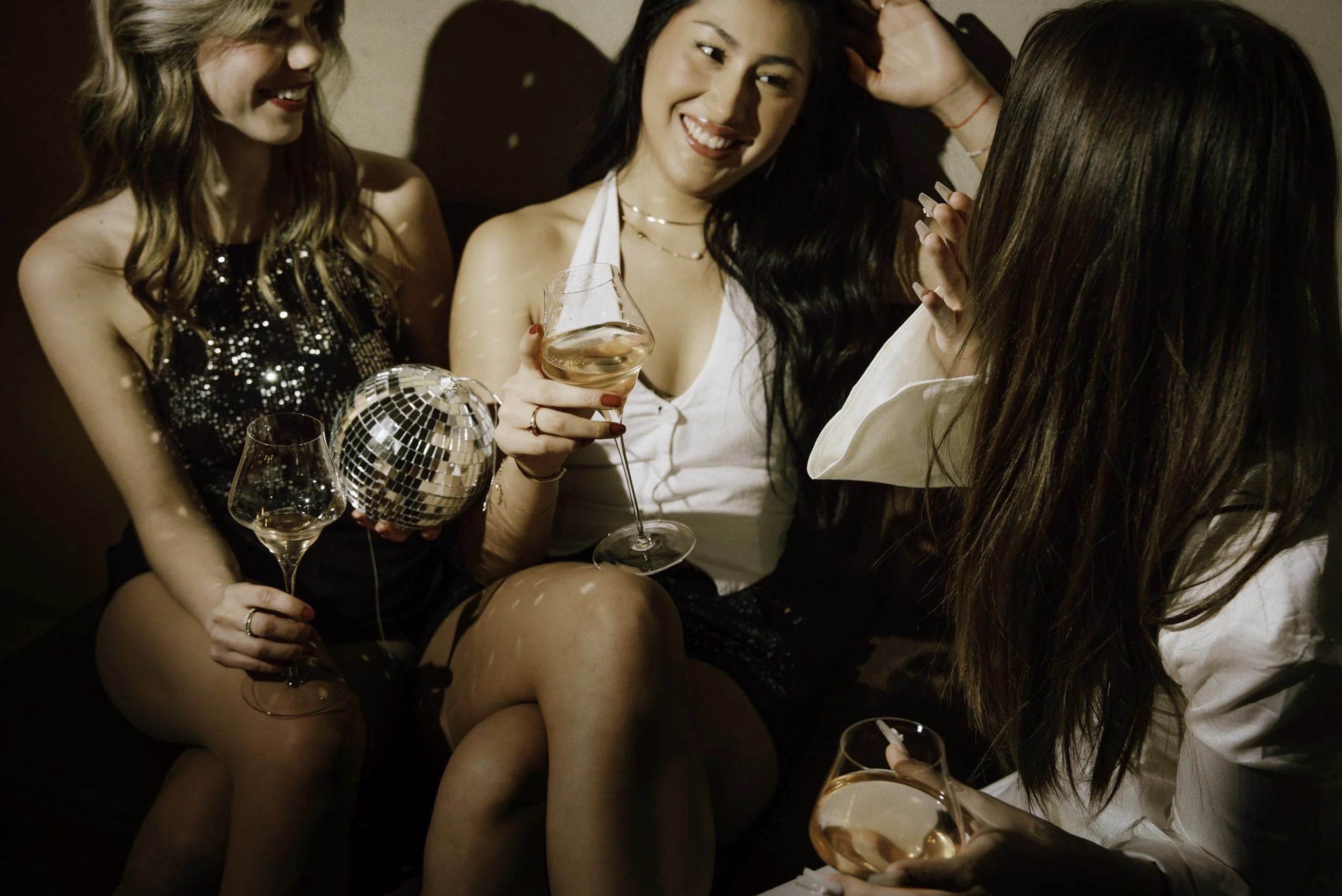 Three women at a party, two of them holding glasses of pink wine and one holding a disco ball, sitting close together on a sofa, smiling and talking.