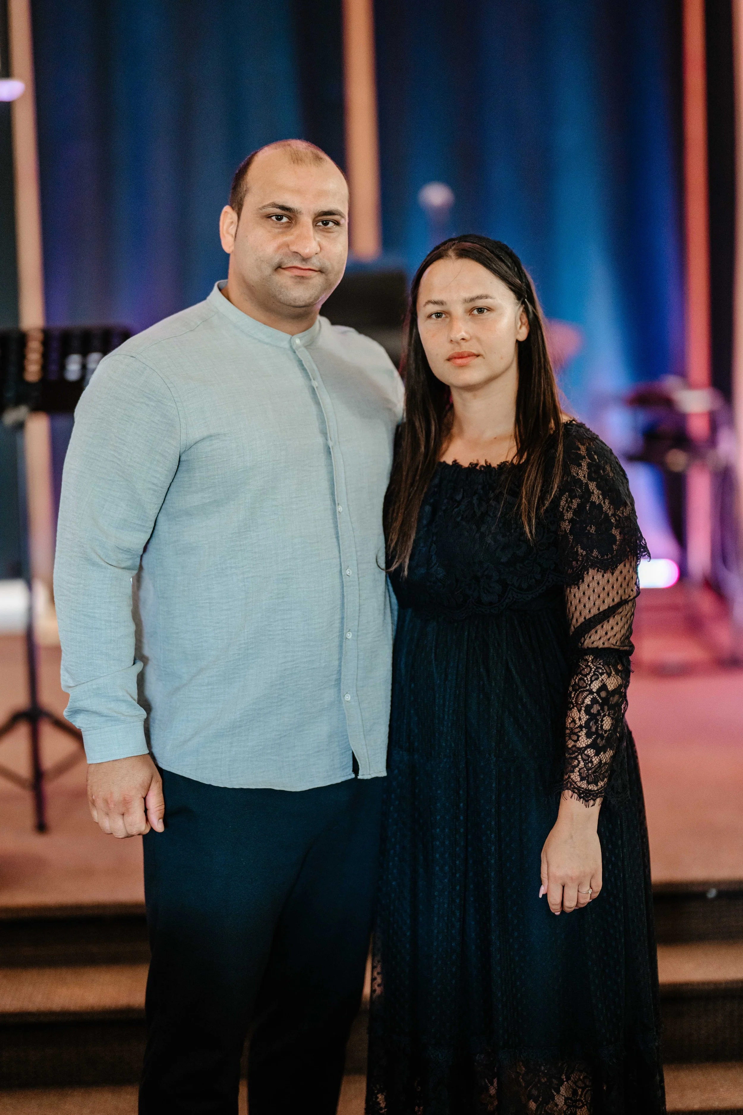 Smiling woman with shoulder-length black hair, wearing a blue top, in a greenhouse with green plants.