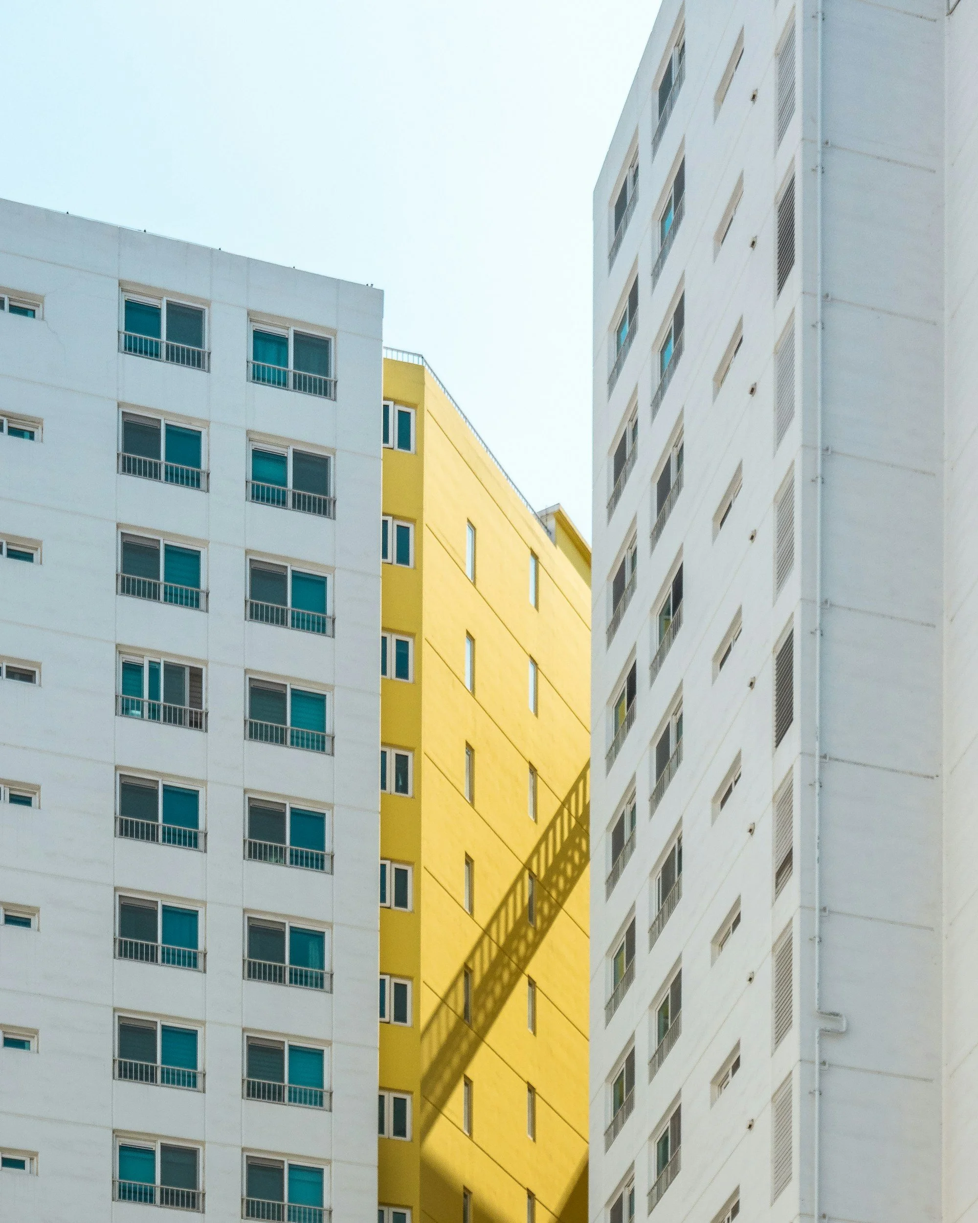 Close-up of white, yellow, and gray modern apartment building facades with windows, some with curtains, under a clear sky. A shadow of a building's balcony railing is cast on the yellow building.