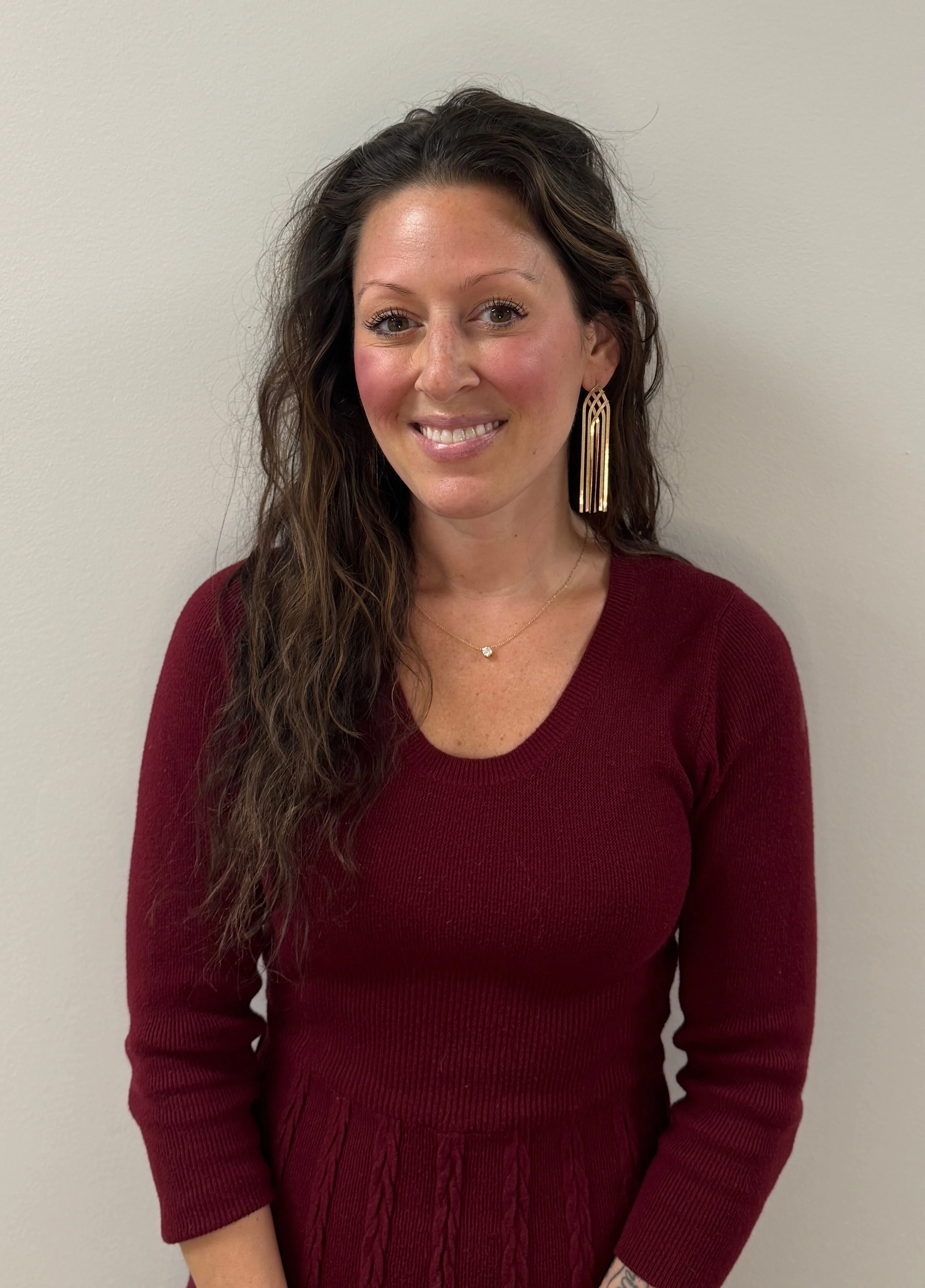 A woman with long wavy brown hair, wearing a burgundy dress and long gold earrings, standing against a plain white wall and smiling.
