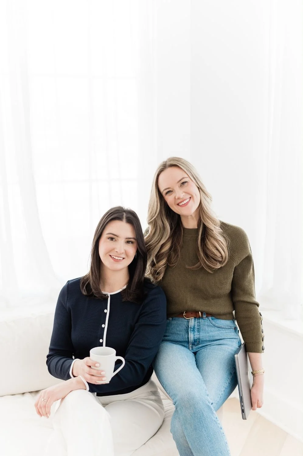 Two women with long hair smiling and sitting together on a white couch in a bright room, one holding a white mug and the other holding a closed laptop.