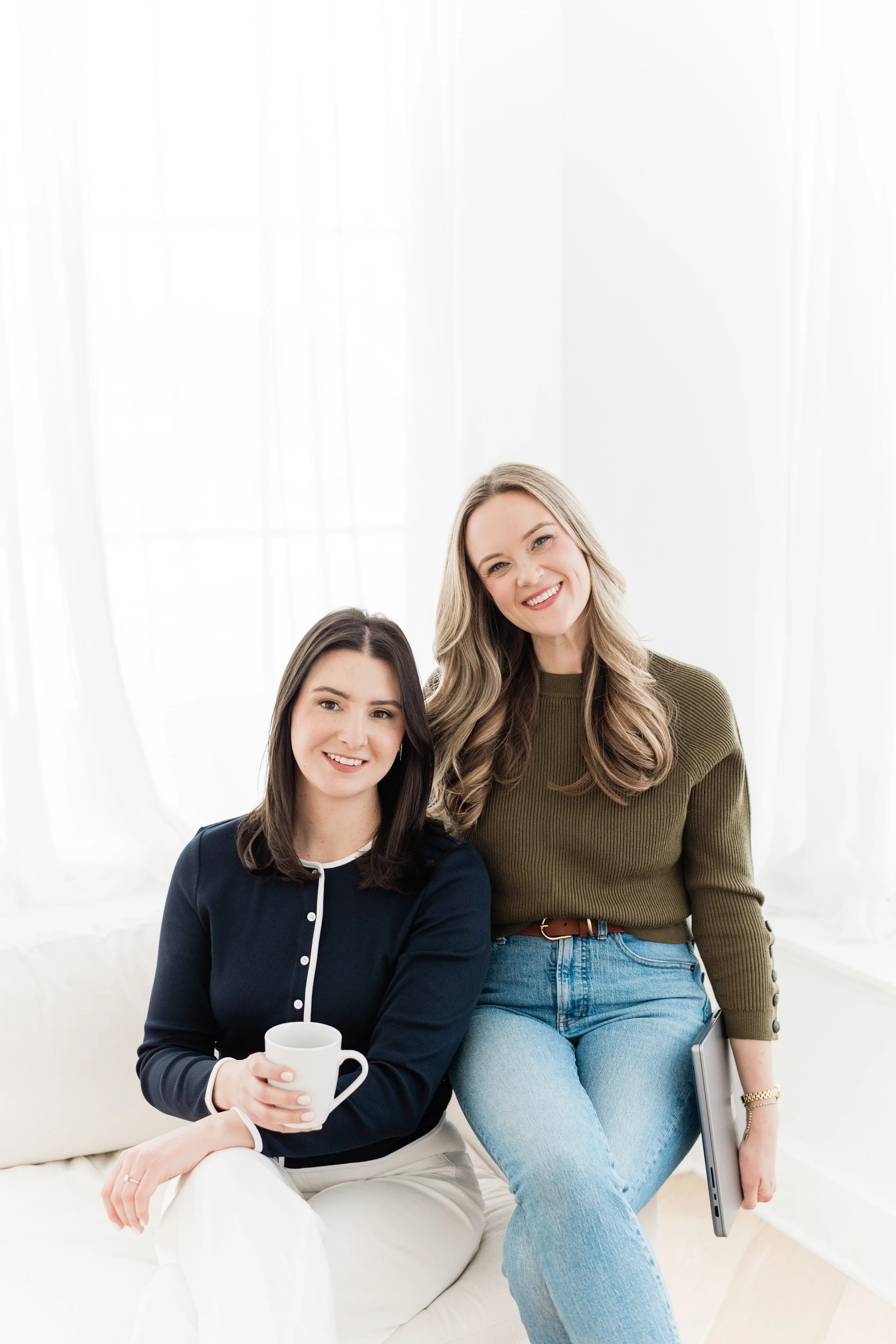 Two women sitting together on a white sofa, smiling at the camera in a bright room with white curtains.