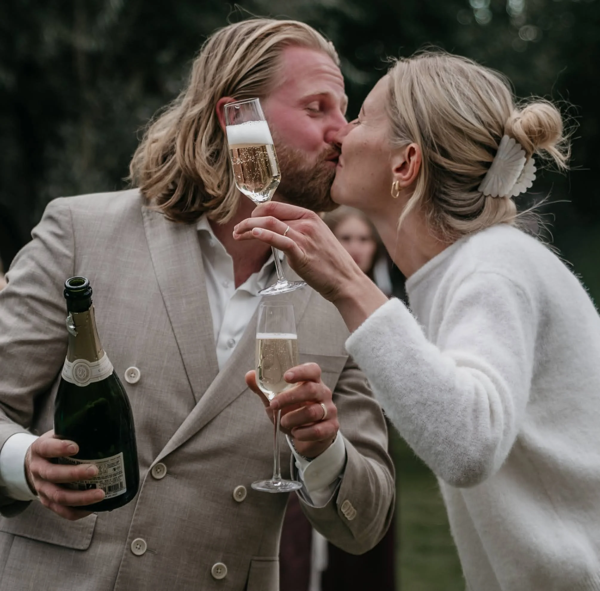 Twee mensen kussen elkaar op een feestje, de man houdt een glas champagne en een fles in de hand.