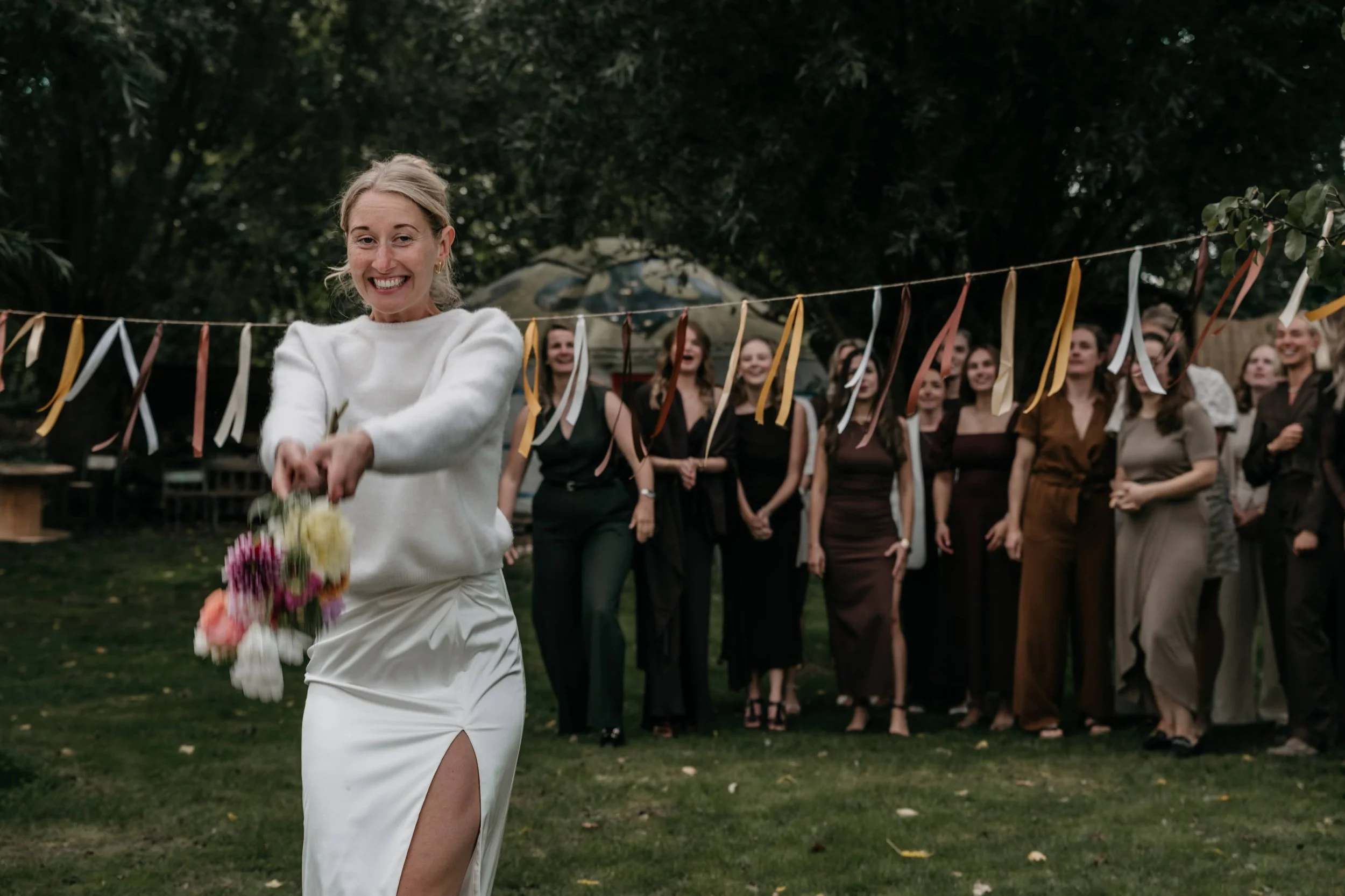 Een vrouw in een witte jurk houdt een bloemenbundel vast en loopt naar een groep vrouwen die lachend en applaudisserend klaarstaan voor een buitenbruiloft. Er hangen kleurrijke linten in de boom.