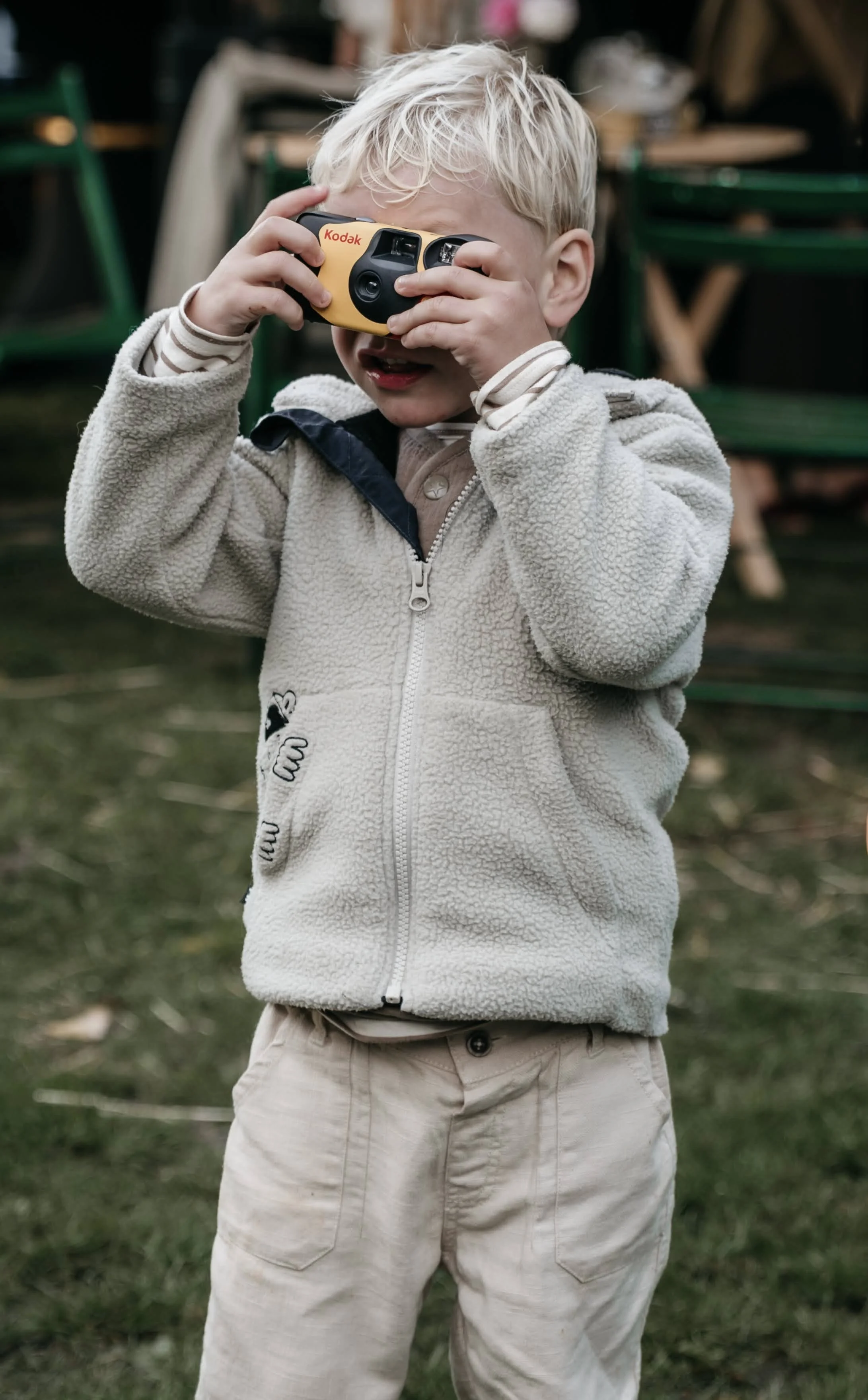 Een jong kind met groen haar draagt een beige fleece jas en beige broeken, en houdt een kleine Kodak-camera voor het gezicht terwijl hij buiten op gras speelt.