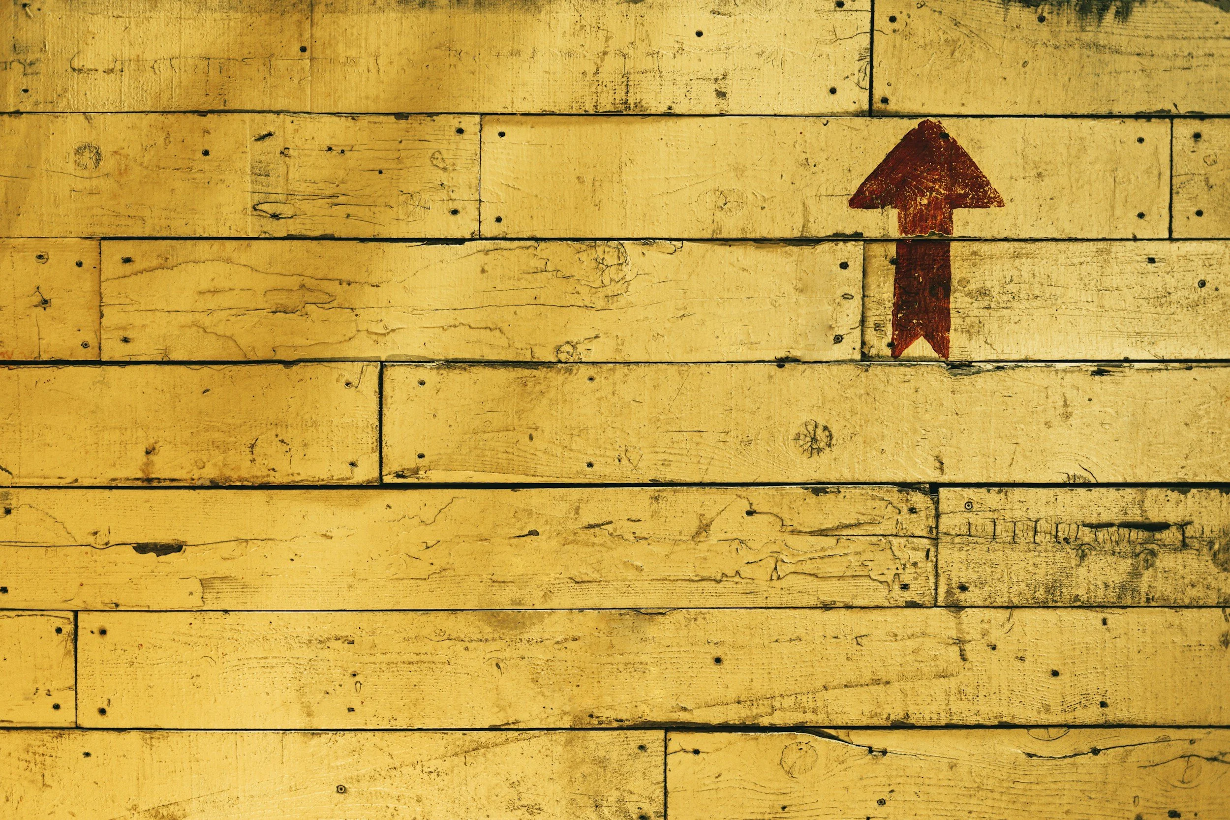 A weathered yellow wooden wall with an upward pointing red arrow painted on it.