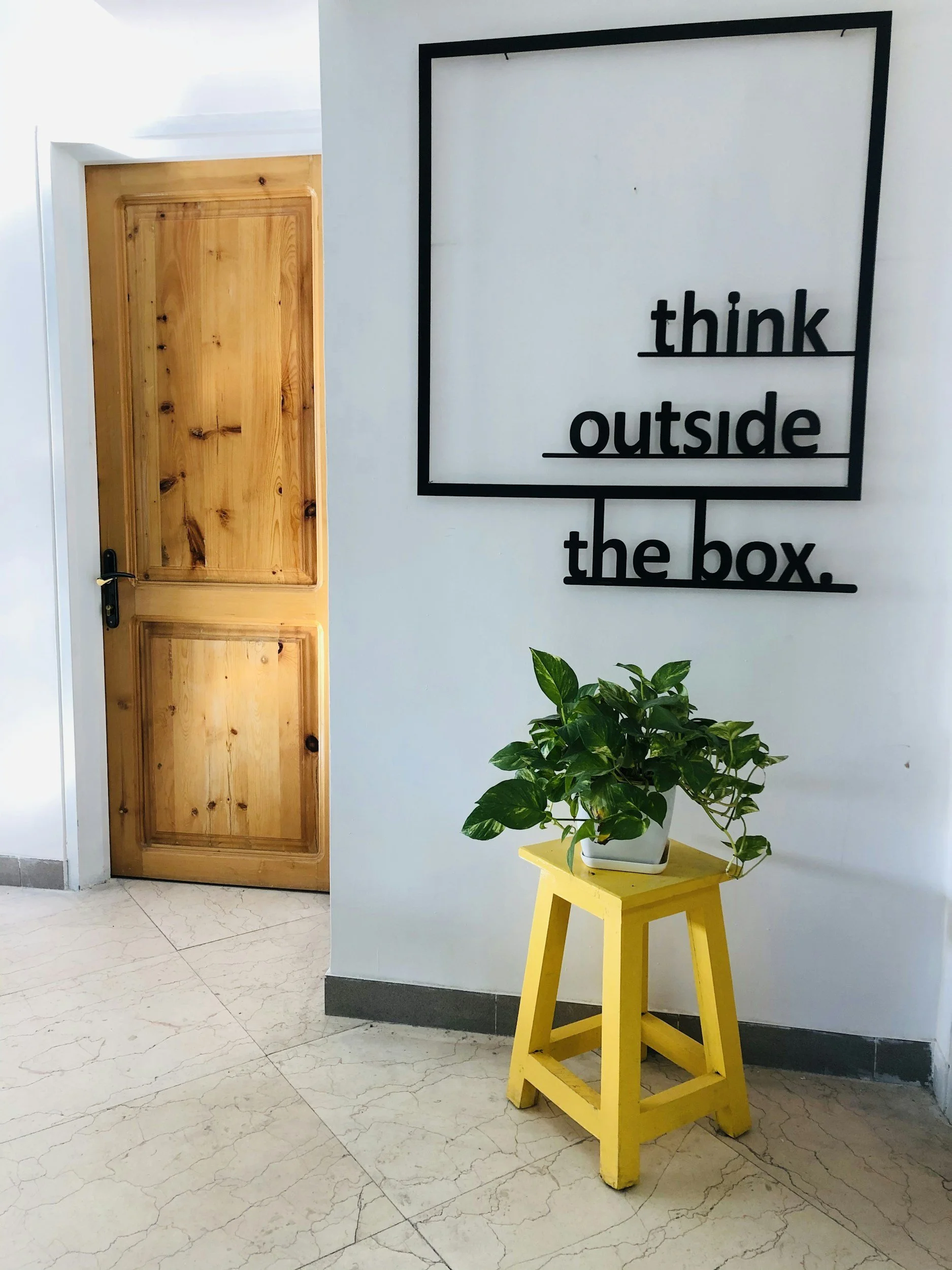 An interior scene with a wooden door on the left, a white wall on the right with a black framed sign reading "think outside the box," a yellow wooden table with a potted green plant, and marble flooring.