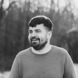 A man with a beard and short hair smiling outdoors in black and white.