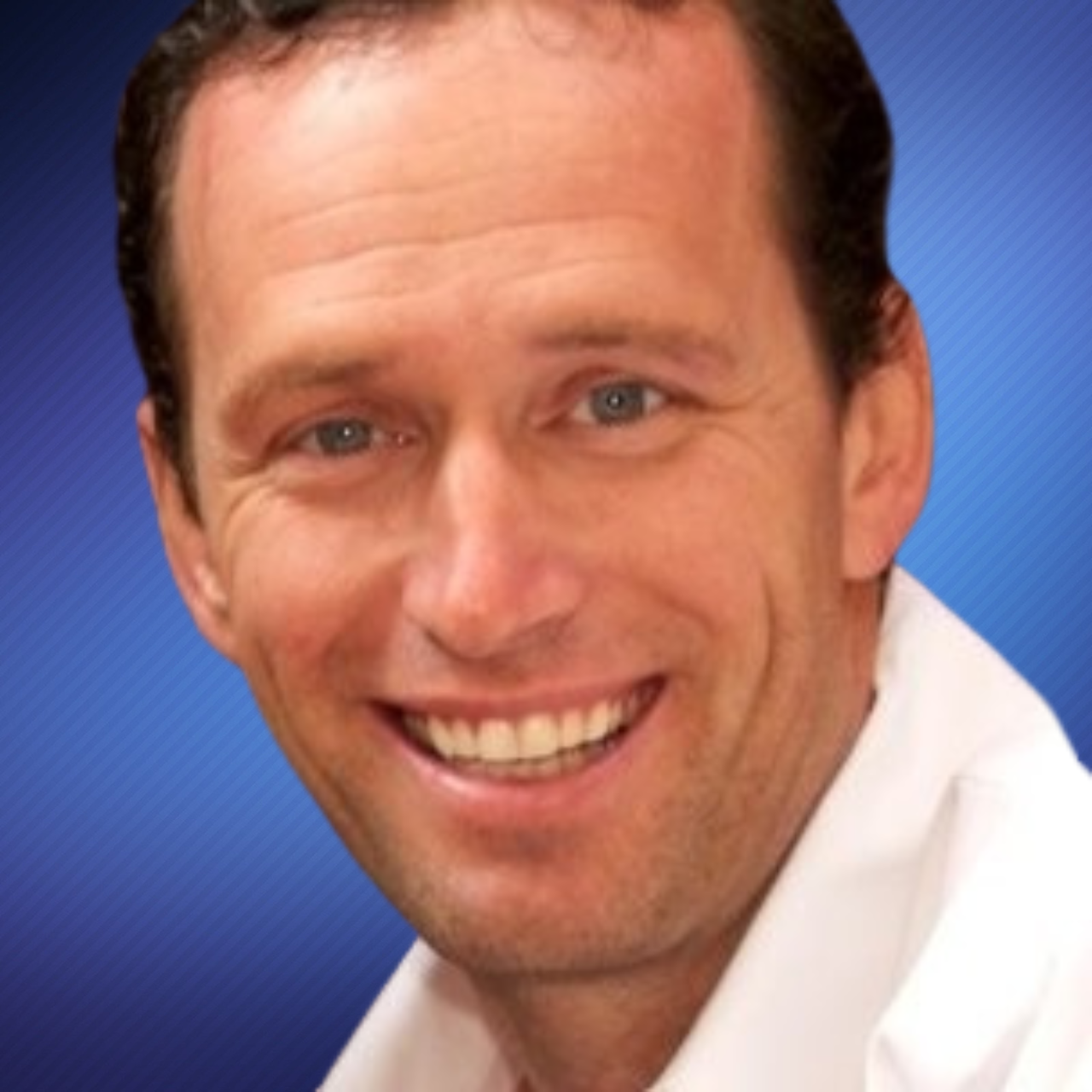 Close-up portrait of a smiling man with light skin, dark brown hair, and blue eyes, wearing a white shirt against a blue background.