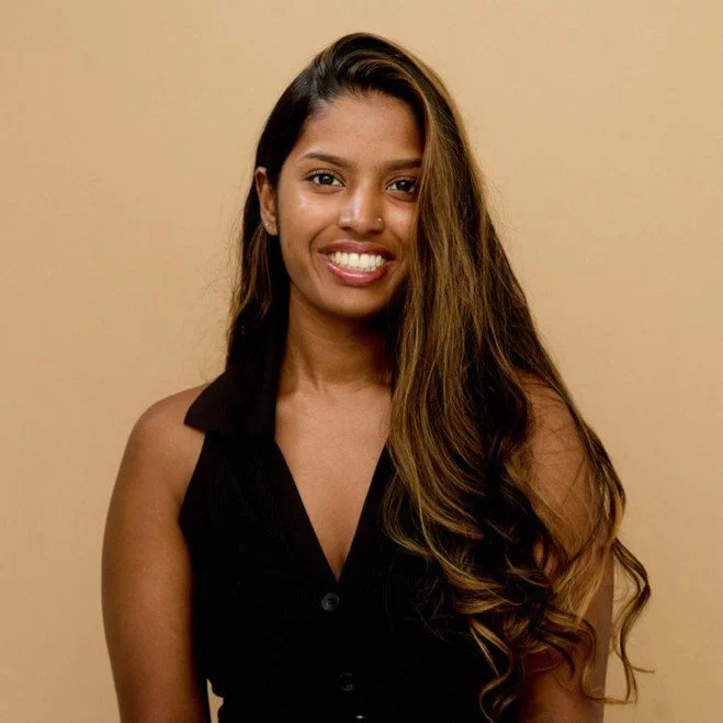 A woman with long, wavy brown hair, wearing a black sleeveless top, smiling against a beige background.