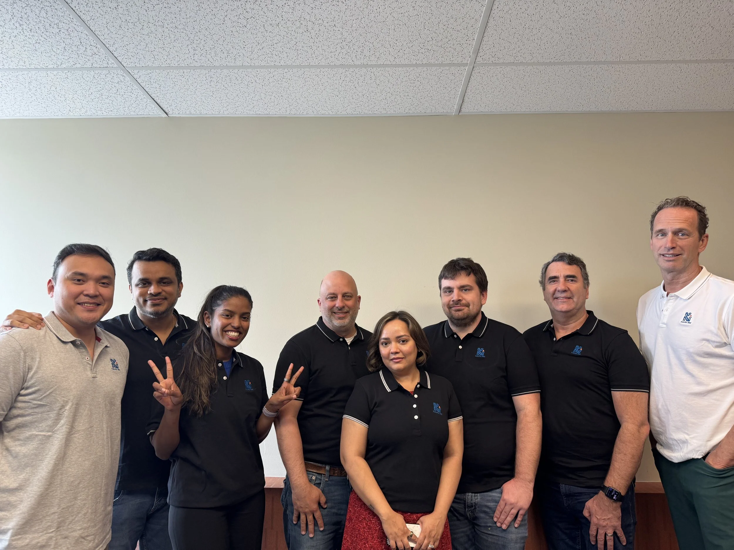 Group of eight diverse people standing in a line against a plain wall, smiling at the camera, some making peace signs.