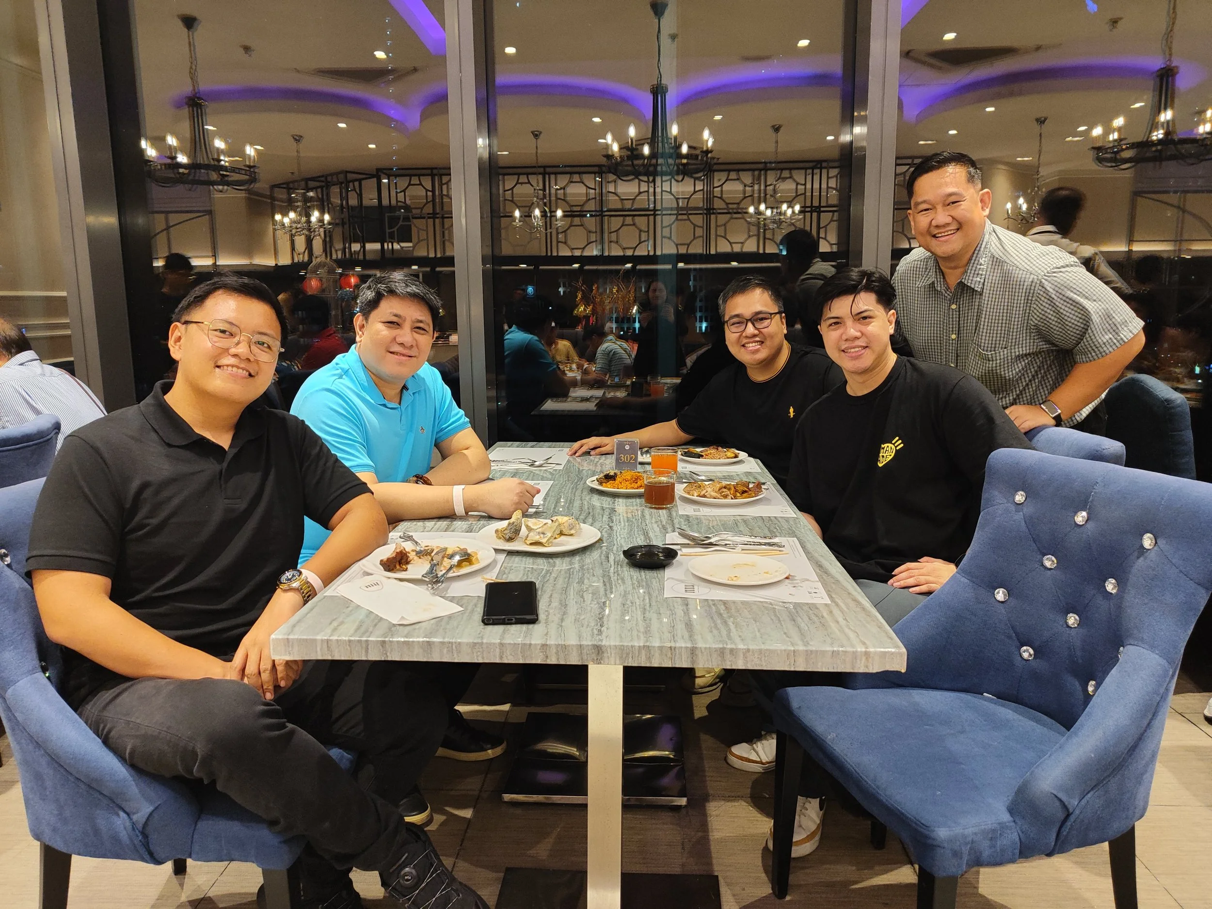 A group of six men sitting and standing around a dining table in a restaurant, smiling and posing for the photo. The table has food and drinks, and the background features large glass windows and elegant lighting.
