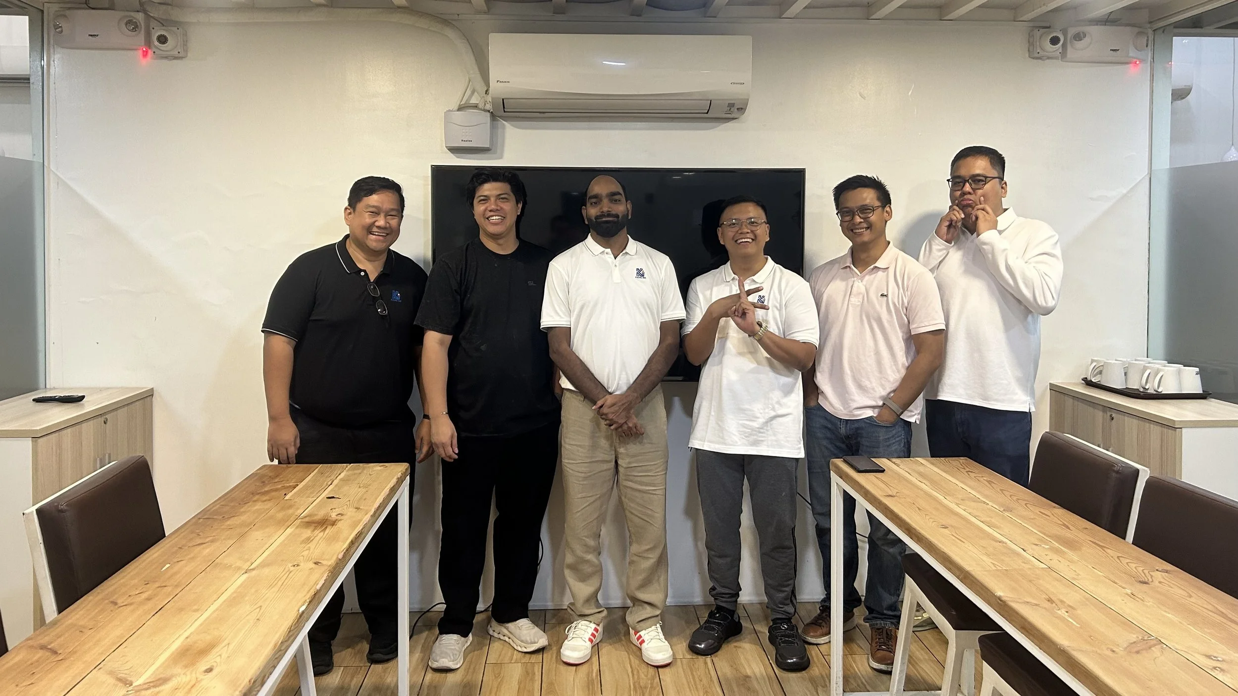 Six men standing in a conference room, smiling at the camera, with wooden tables and a TV screen behind them.