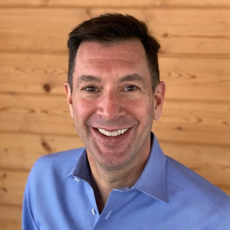 A smiling man with short dark hair wearing a blue dress shirt, standing in front of a wooden wall.