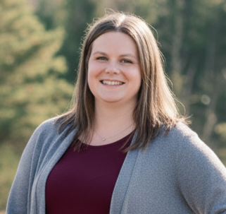 A woman with shoulder-length brown hair smiling outdoors, wearing a gray cardigan over a burgundy top, with trees in the background.