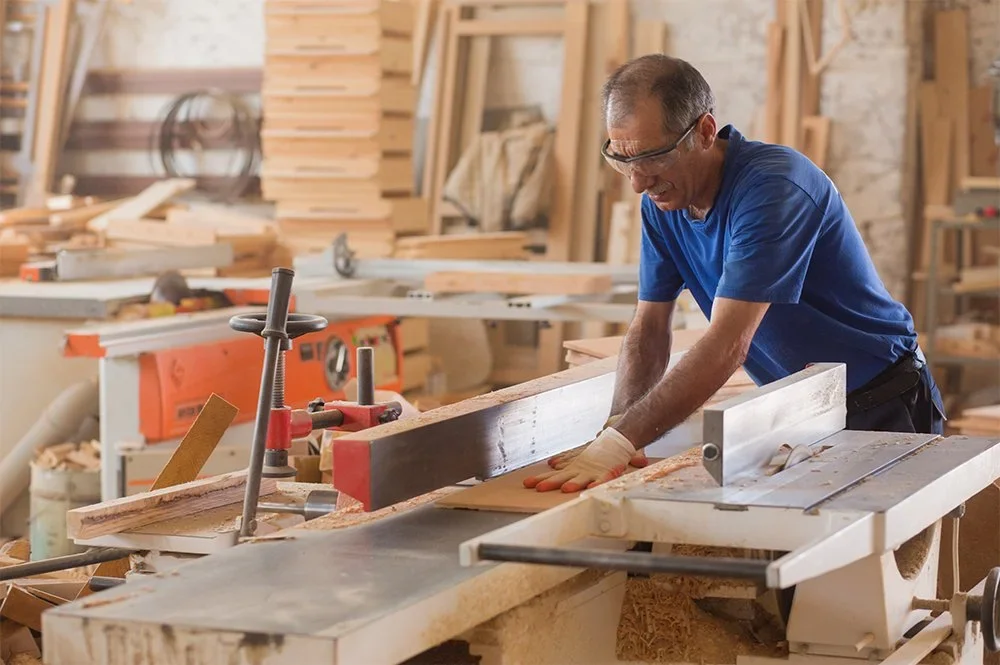 An elderly man wearing safety glasses and gloves working with wood at a saw in a woodworking shop.
