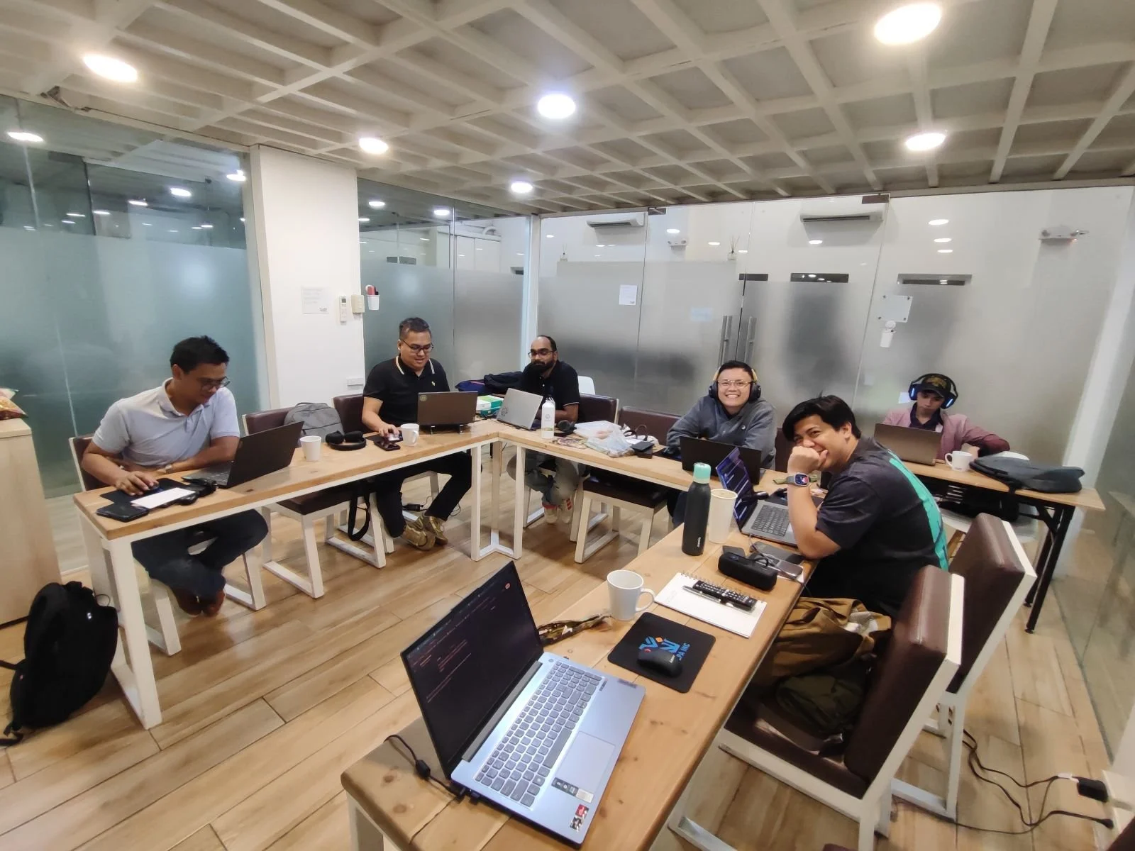 Group of six people sitting around a conference table with laptops in a modern office meeting room, some smiling and looking at the camera.