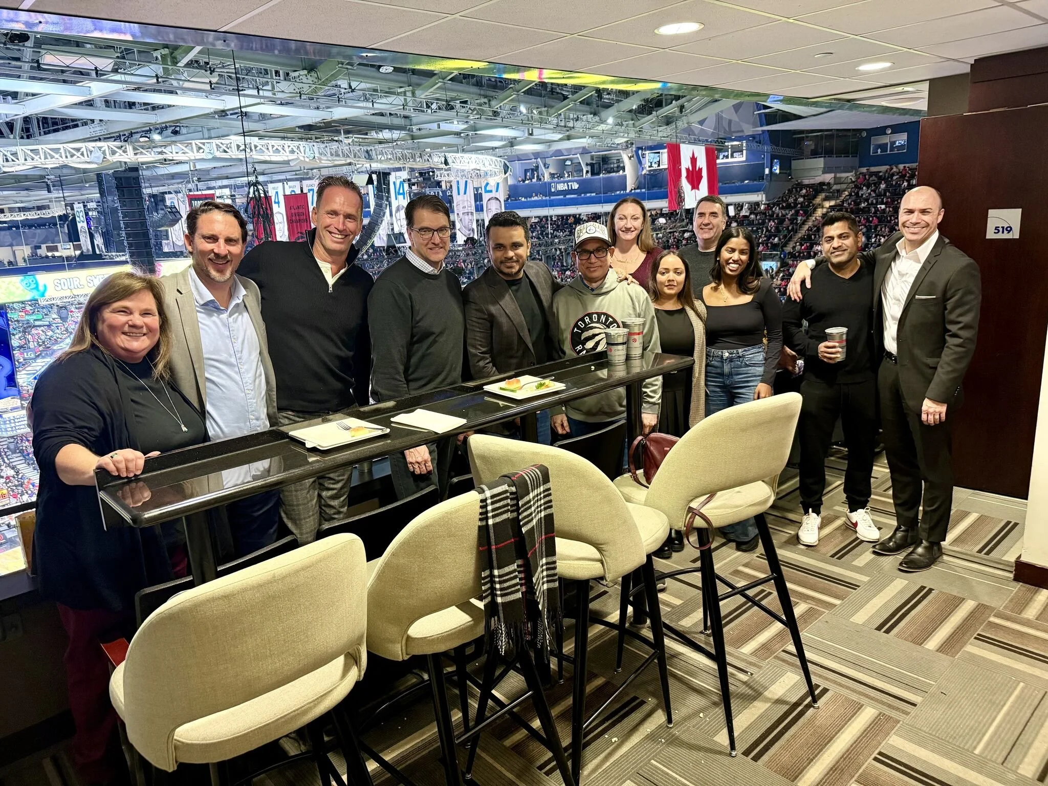Group of eleven people standing by a bar counter inside a sports arena, with a hockey game visible in the background, some holding coffee cups.