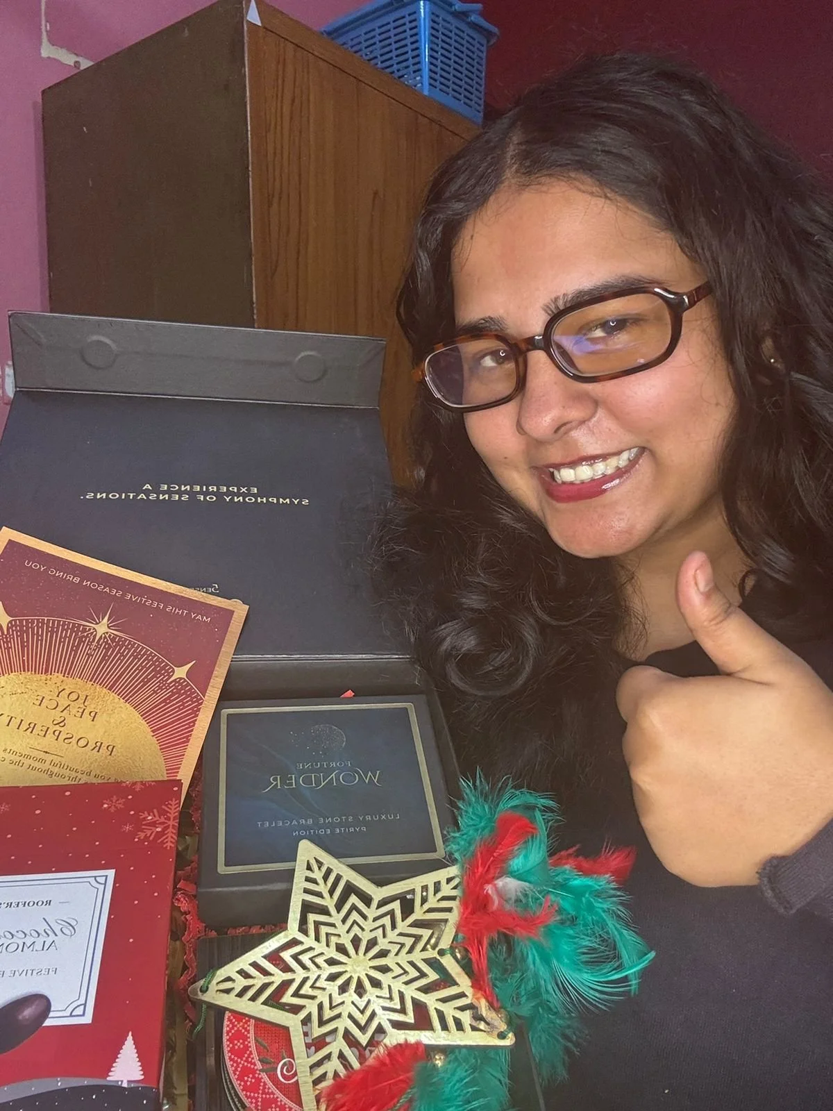 A woman with dark curly hair and glasses smiling and giving a thumbs up next to a holiday gift box, decorations, and a present labeled 'WONDER.'