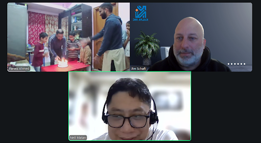 A virtual meeting with three participants on a video call. The top left shows a group of children and two adults gathered around a table with a birthday cake. The top right features a man with a beard and a shaved head in front of a plant and a logo.