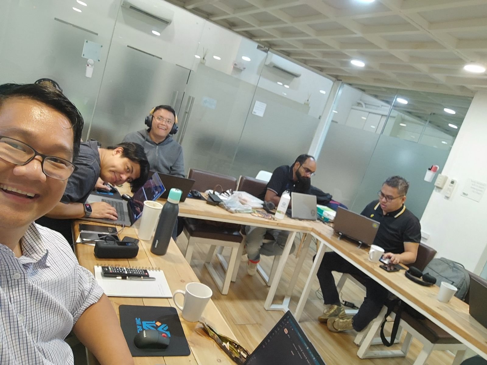 Group of five men working and socializing in a modern office conference room with laptops, notebooks, coffee mugs, and personal items on the table.
