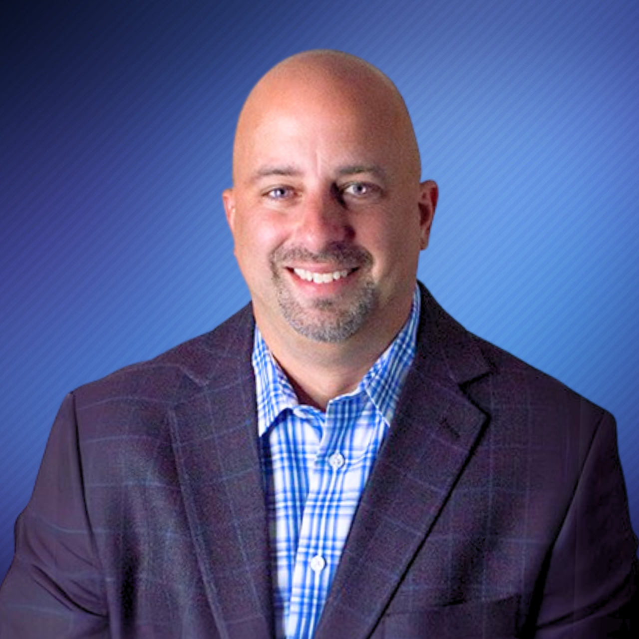 Headshot of a smiling bald man with a goatee, wearing a dark plaid suit and blue checkered shirt, against a blue striped background.