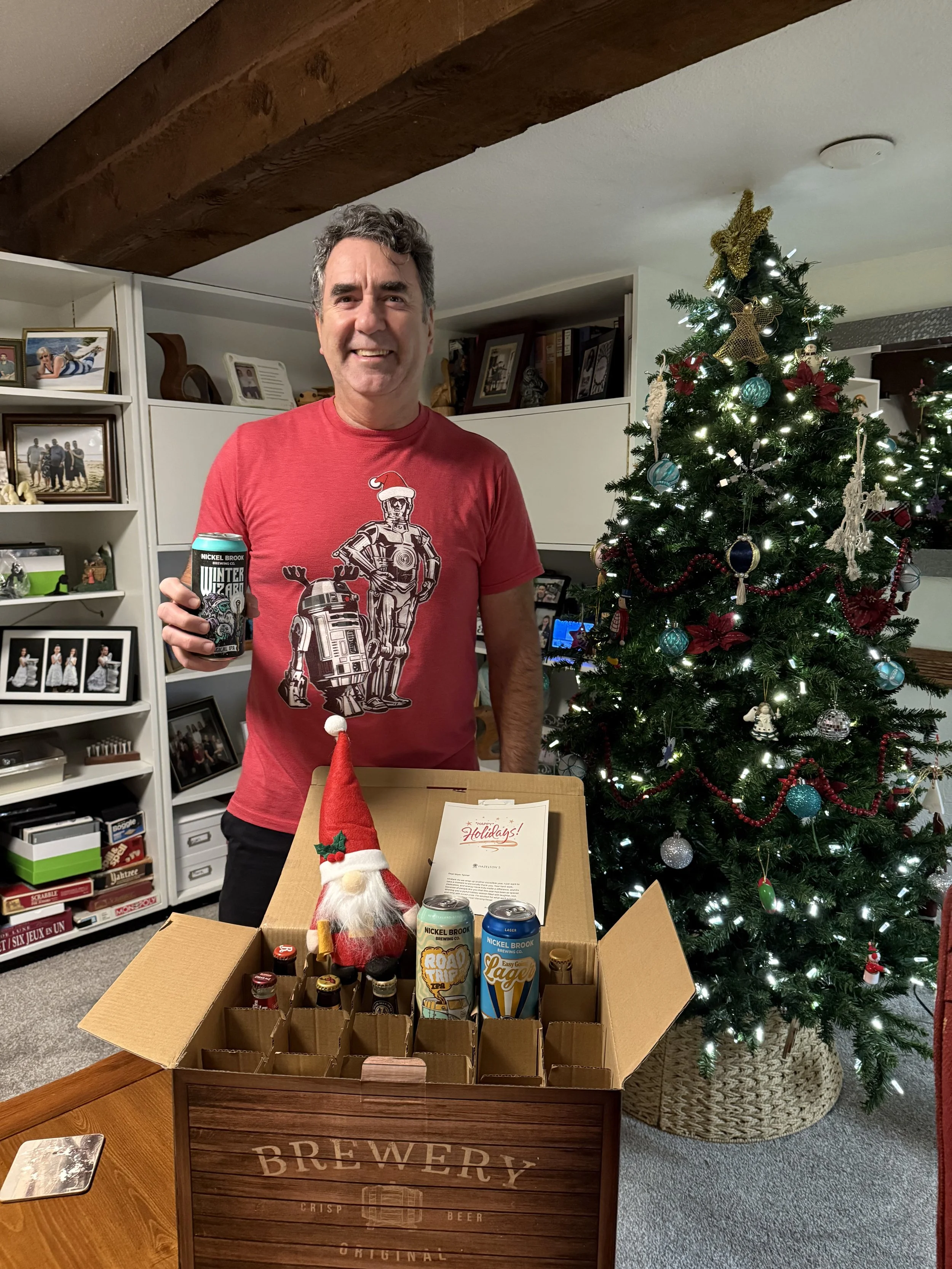 A man standing next to a decorated Christmas tree, holding a can of beer, with a gift box containing various beverages and a Santa figure on a table.