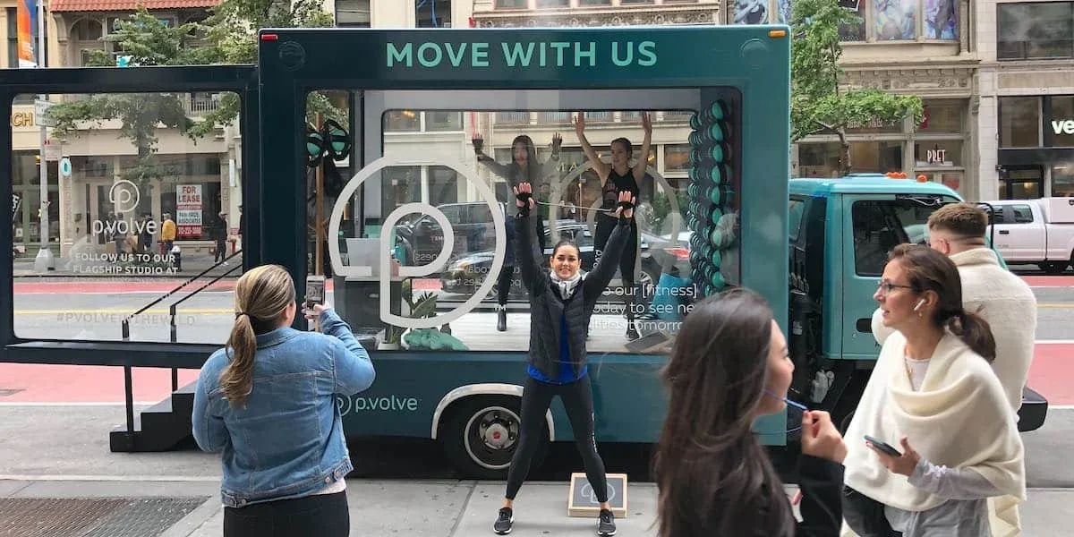 A group of people is gathered in front of a teal-colored mobile fitness studio truck with the words 'MOVE WITH US' on top. Inside the truck, two women are exercising with resistance bands, and one woman in the foreground is posing with her arms raised, smiling at the camera. Others are taking photos or talking nearby on a city street.