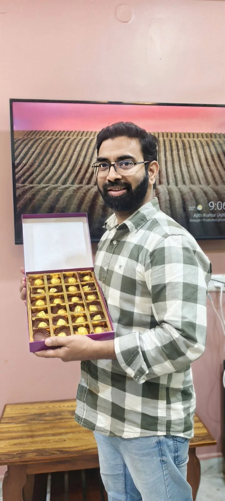 A man in a green and white plaid shirt holding an open box of assorted chocolates, standing in front of a television screen showing a landscape of fields at sunset, inside a room with pink walls and a wooden table.
