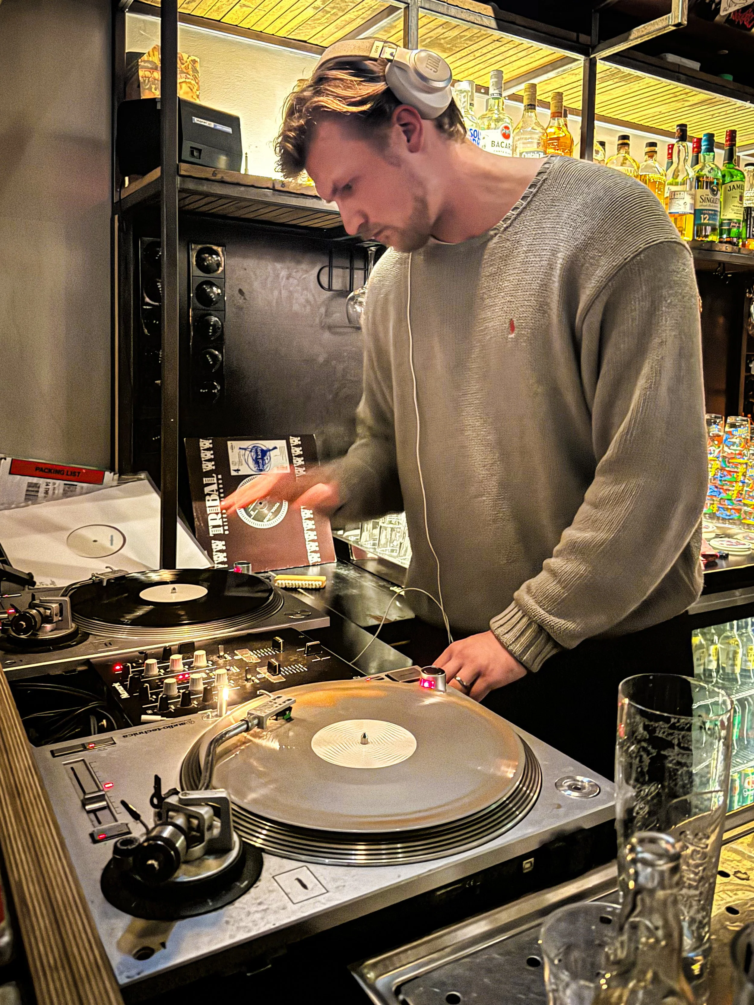 A male DJ in a beige sweater is mixing music on turntables in a bar setting, wearing headphones with one ear covered.