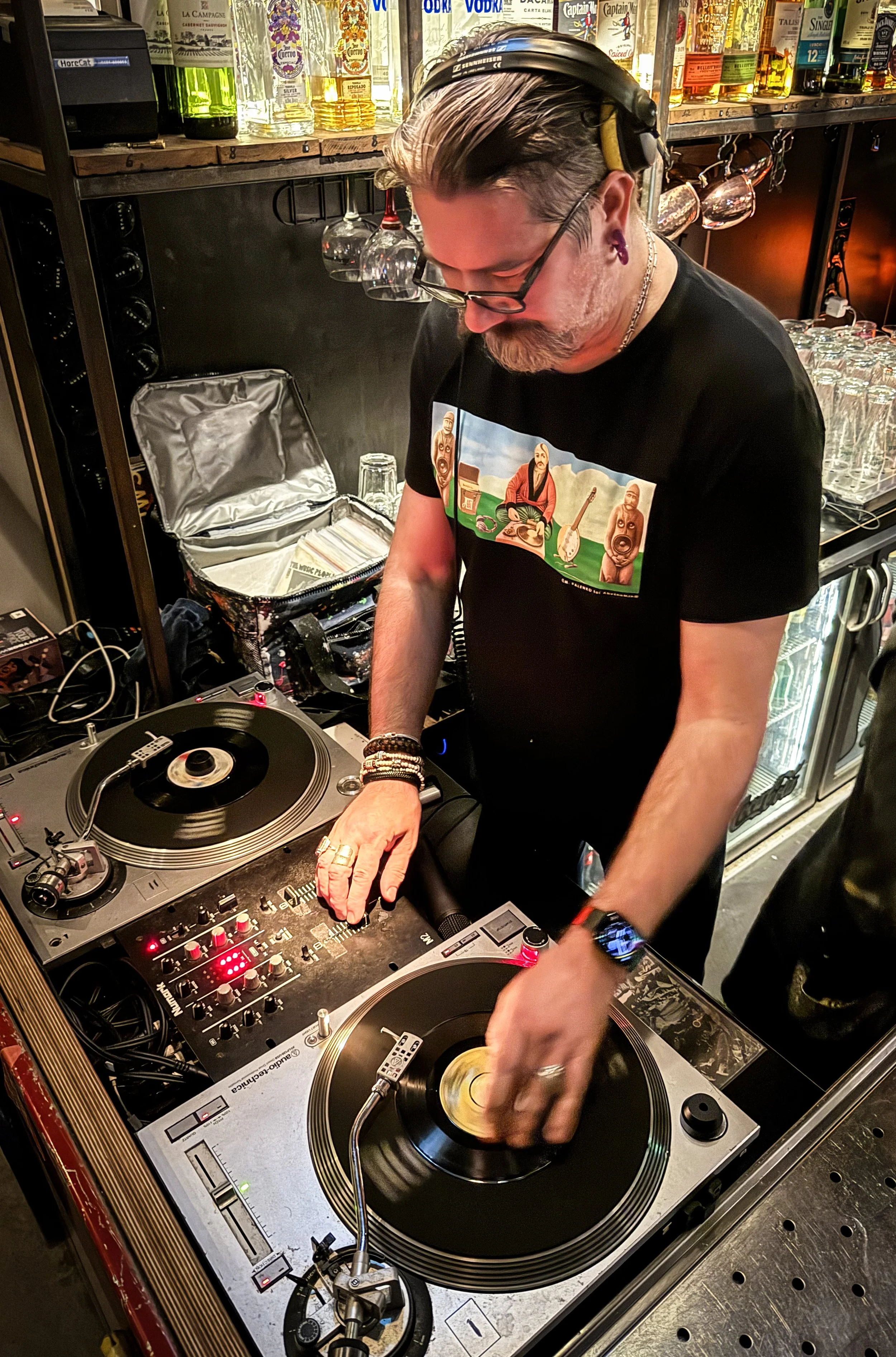 A DJ with black hair and glasses performing with turntables in a bar or club, with liquor bottles and glasses in the background.