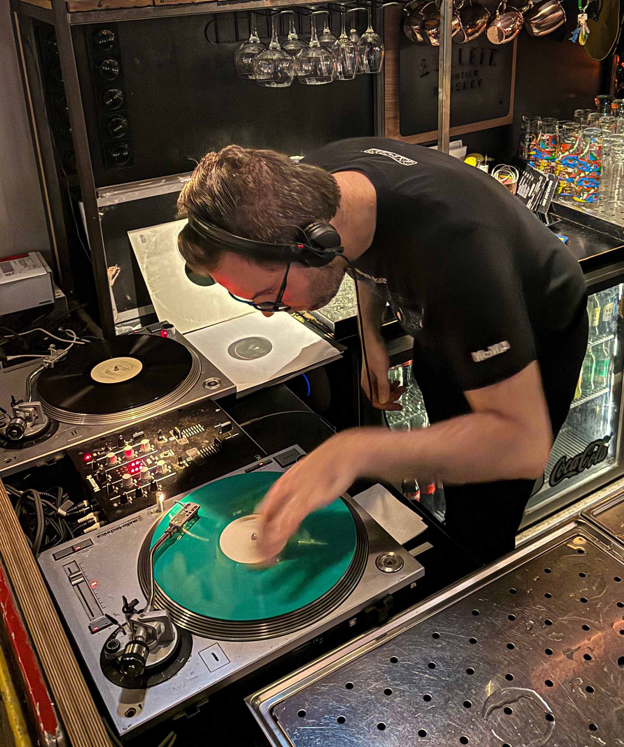 A DJ wearing headphones is operating DJ equipment, including turntables with green vinyl records and a mixer, in a bar or club setting with glasses and mugs hanging above the counter.
