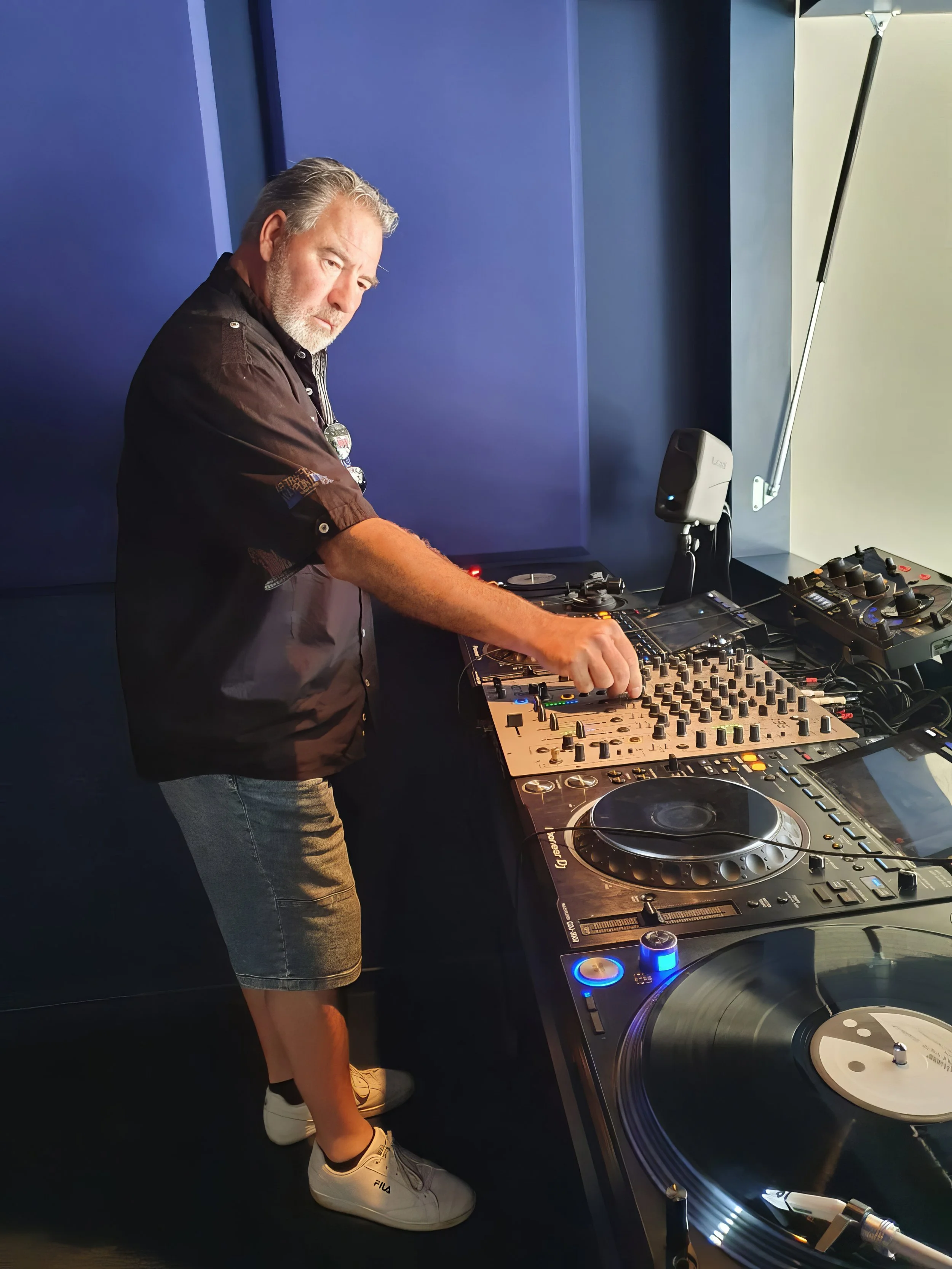 A man with gray hair and a beard DJing in a room with blue walls, wearing a black shirt, gray shorts, and white sneakers, operating DJ equipment.