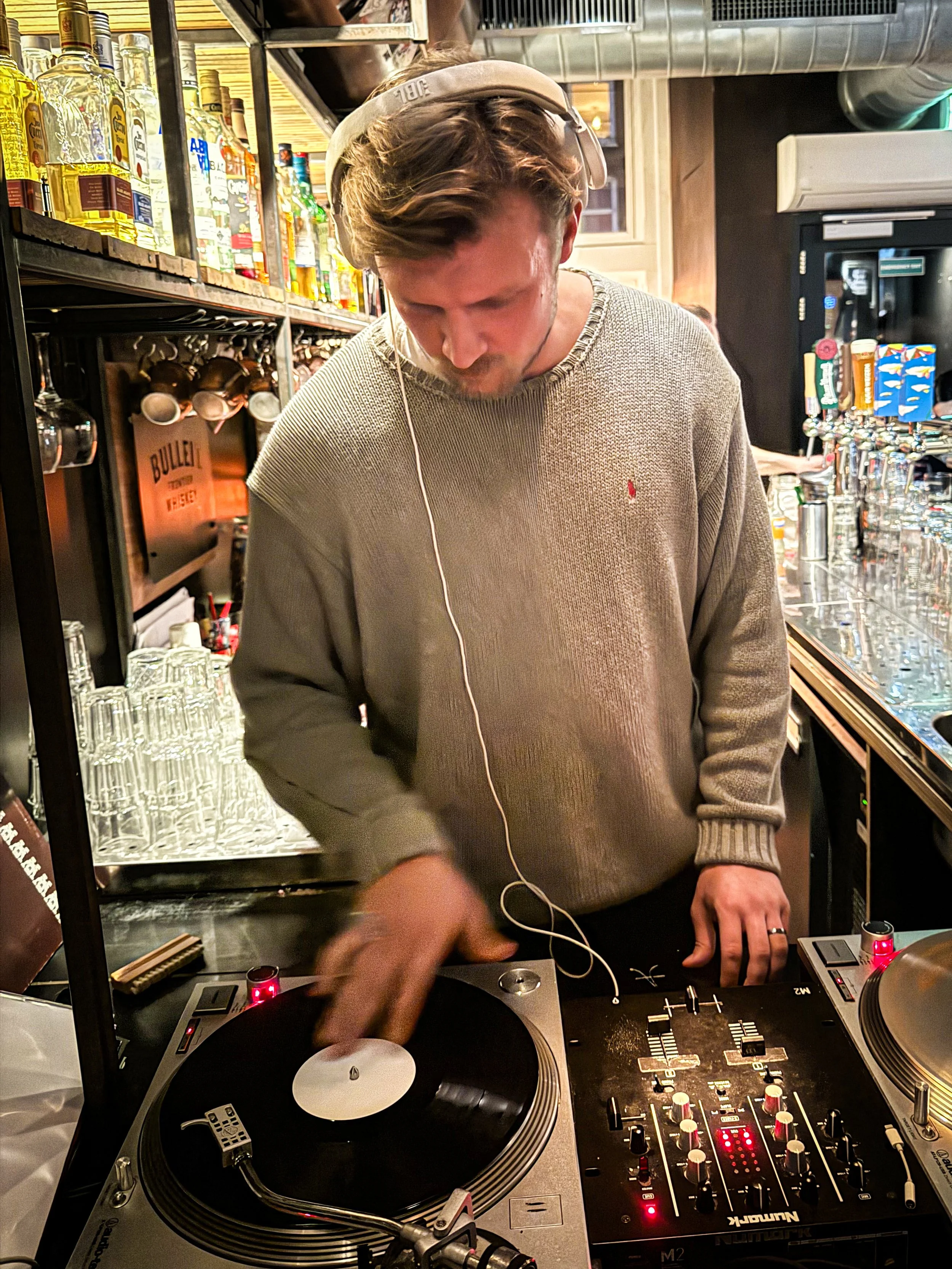 A man DJing in a bar, wearing a gray sweater, headphones, and controlling turntables and a mixer.