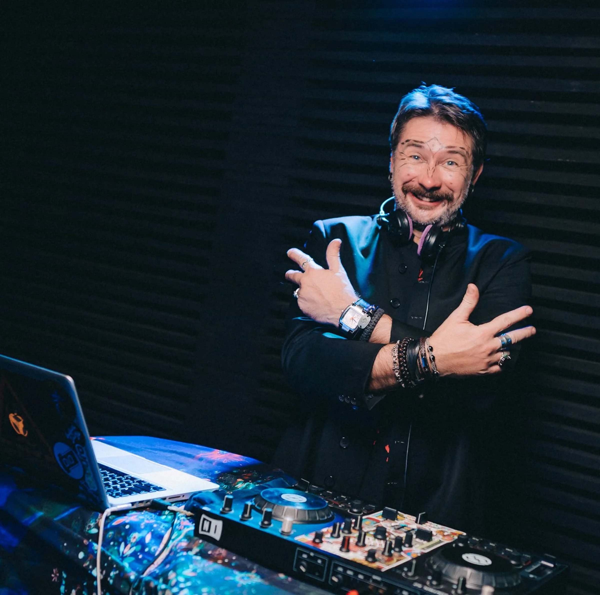 DJ Necto Lucas, a man with a beard and tattoos, smiling and pointing while standing behind a DJ setup with laptop, turntables, and mixer in a dark room.