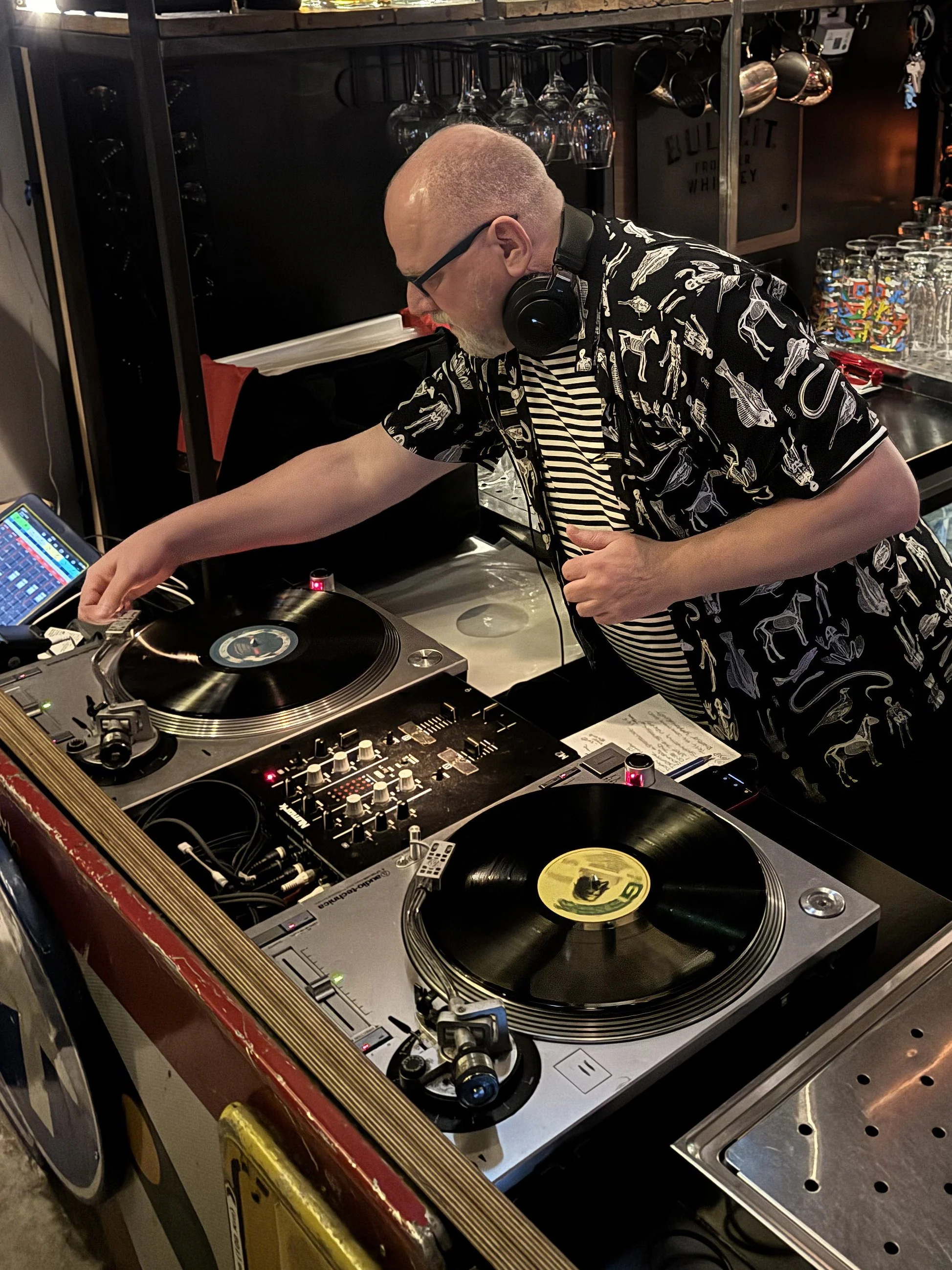 A DJ wearing glasses, headphones, and a patterned shirt, adjusts equipment at a nightclub or bar, with drinks and supplies in the background.