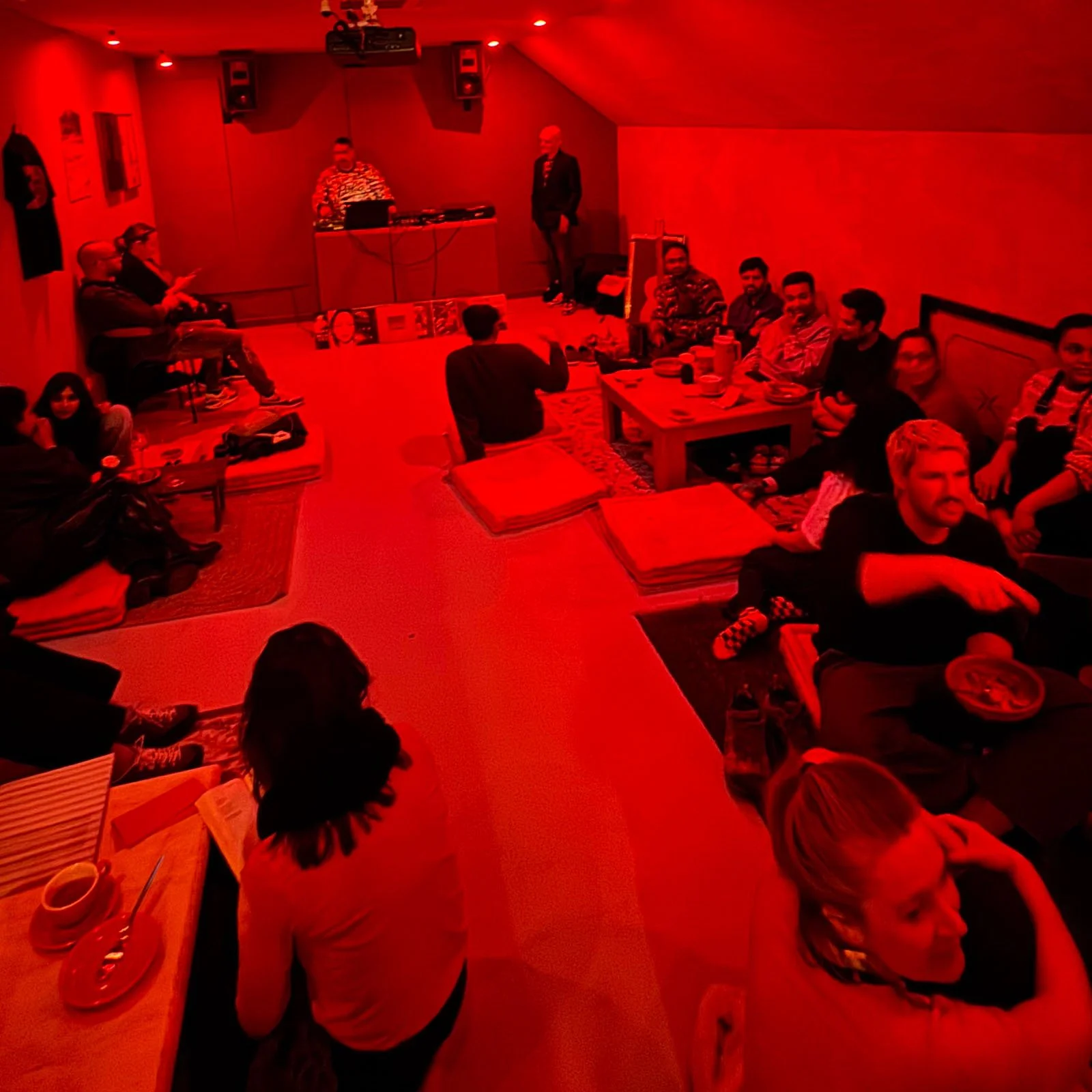 People gathered in a dimly lit room with red lighting, sitting on the floor and chairs, during an an Amsterdam Vinyl Club live event.