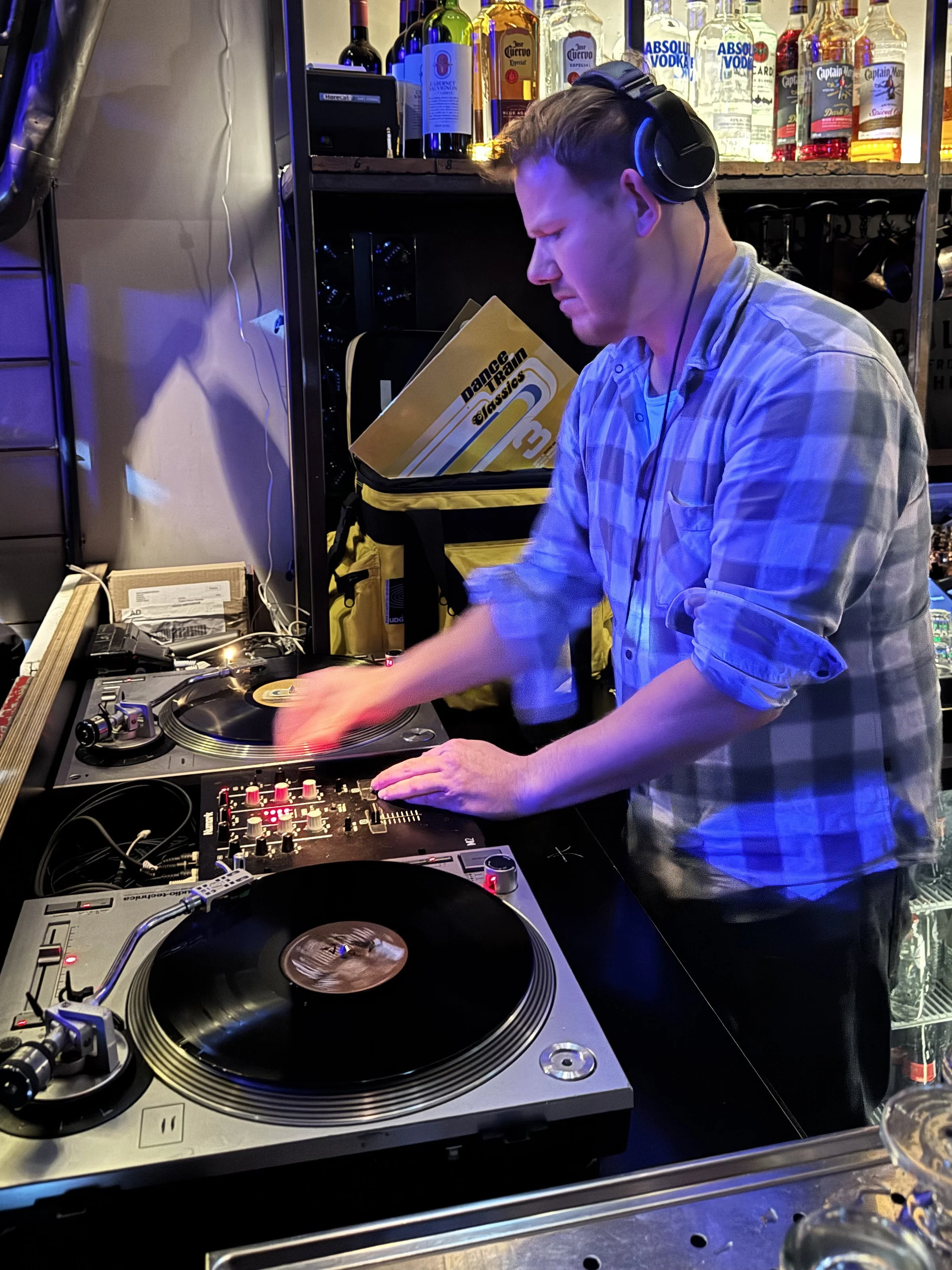 Walter Vooys, wearing headphones and a blue checked shirt, plays a live DJ set for Amsterdam Vinyl Club at the venue Vinyl Rocks in Amsterdam. Liquor bottles can be seen on the shelves behind him.