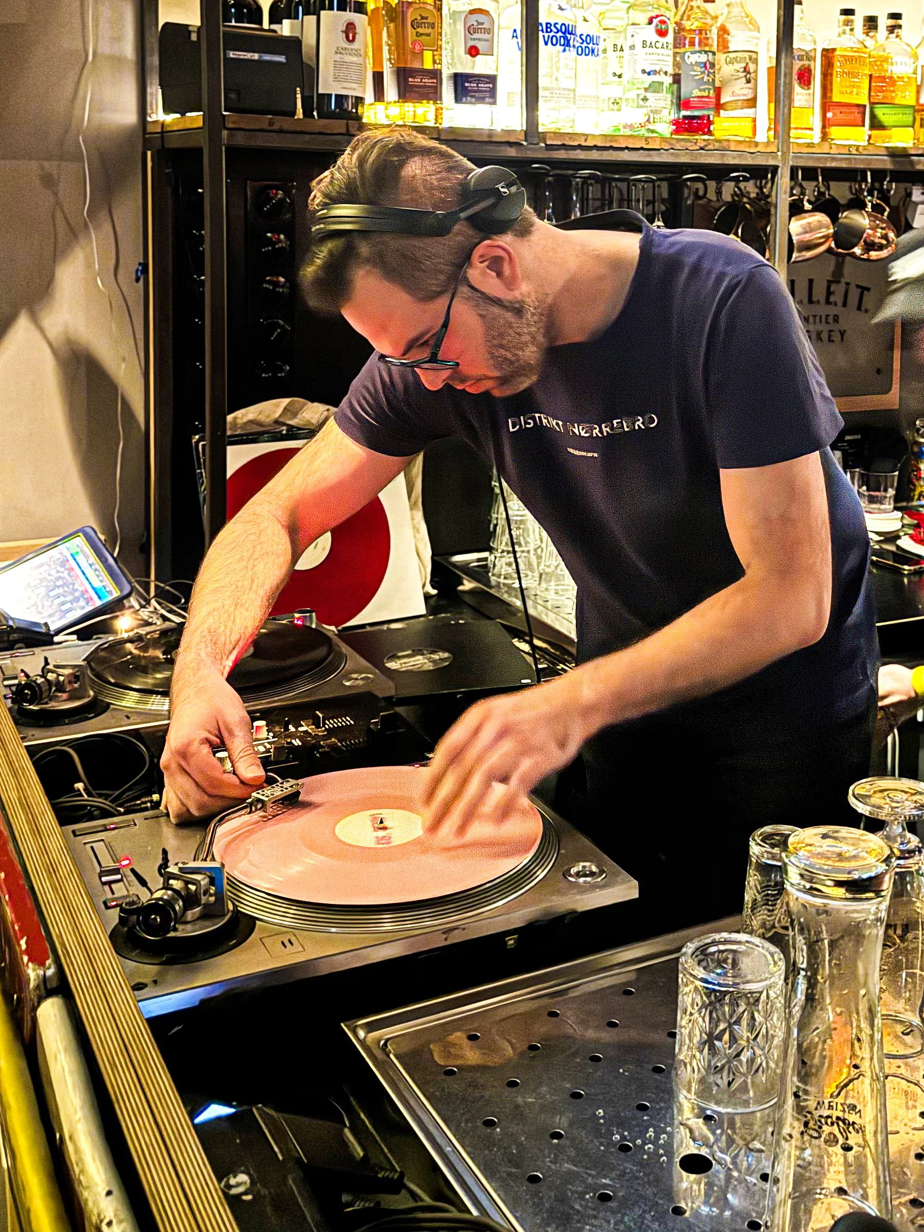 DJ playing music on turntables in a bar with liquor bottles on shelves behind him.