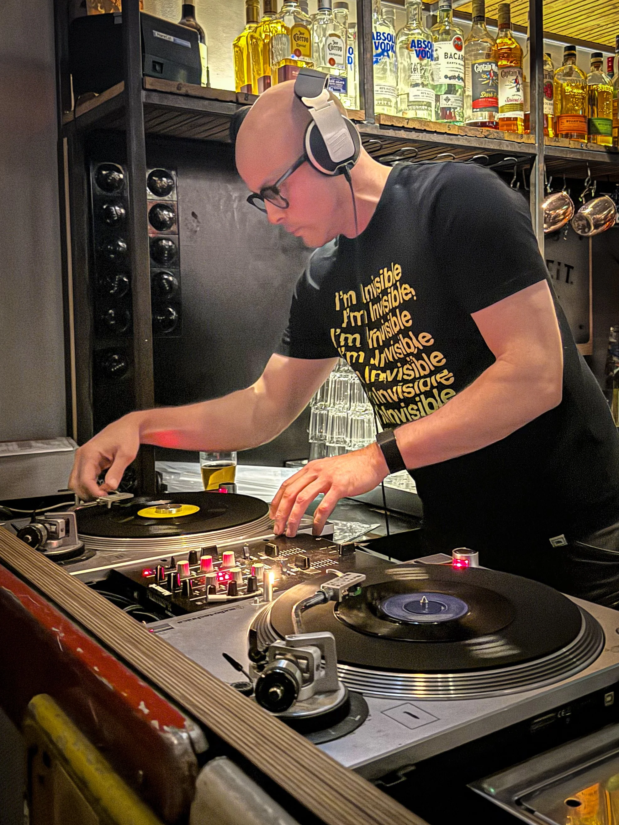 A DJ wearing glasses, headphones, and a black t-shirt with yellow text, is mixing music at a bar with bottles of alcohol in the background.