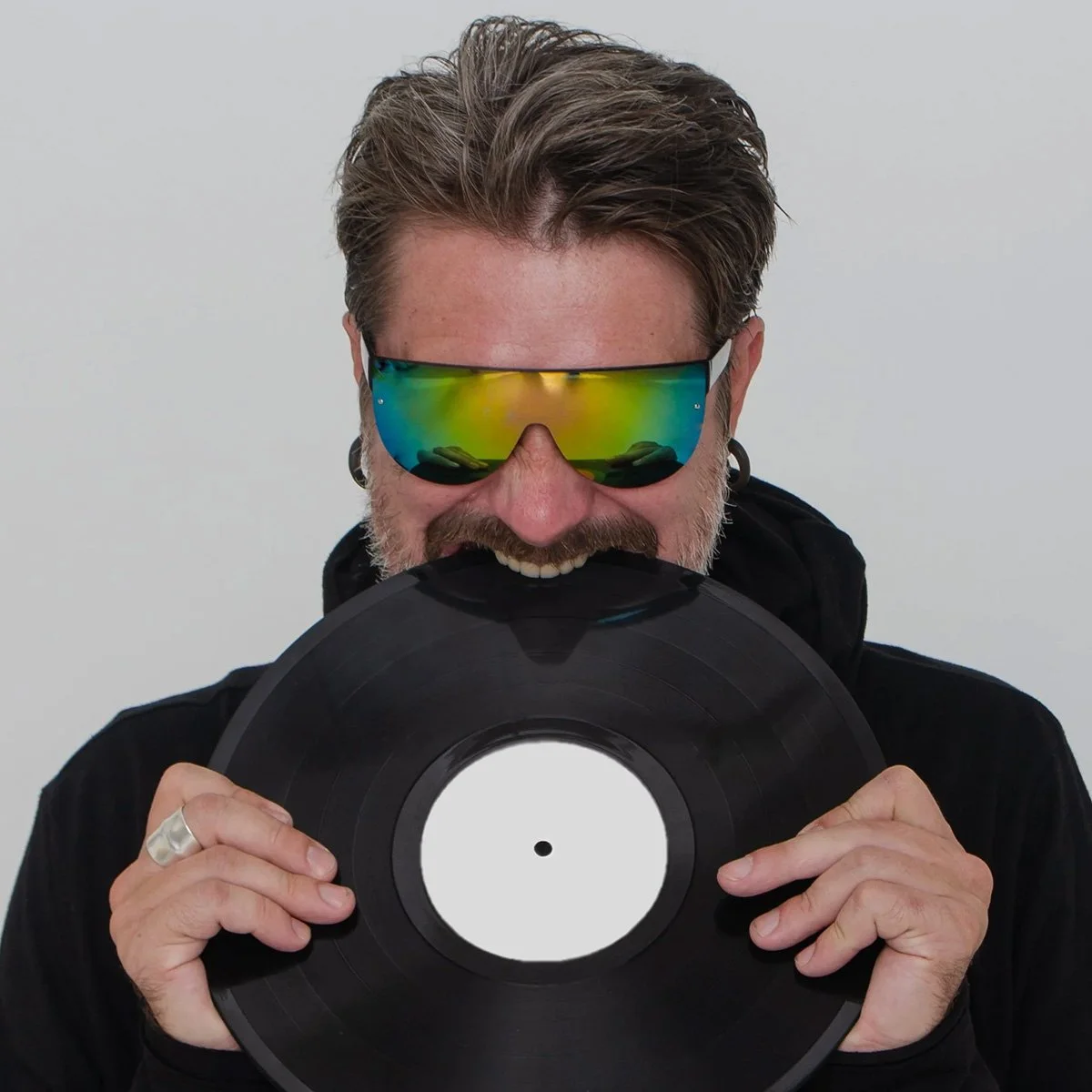 DJ Necto Lucas - a man with gray hair and beard wearing reflective sunglasses holding a vinyl record in front of his face against a plain light background.