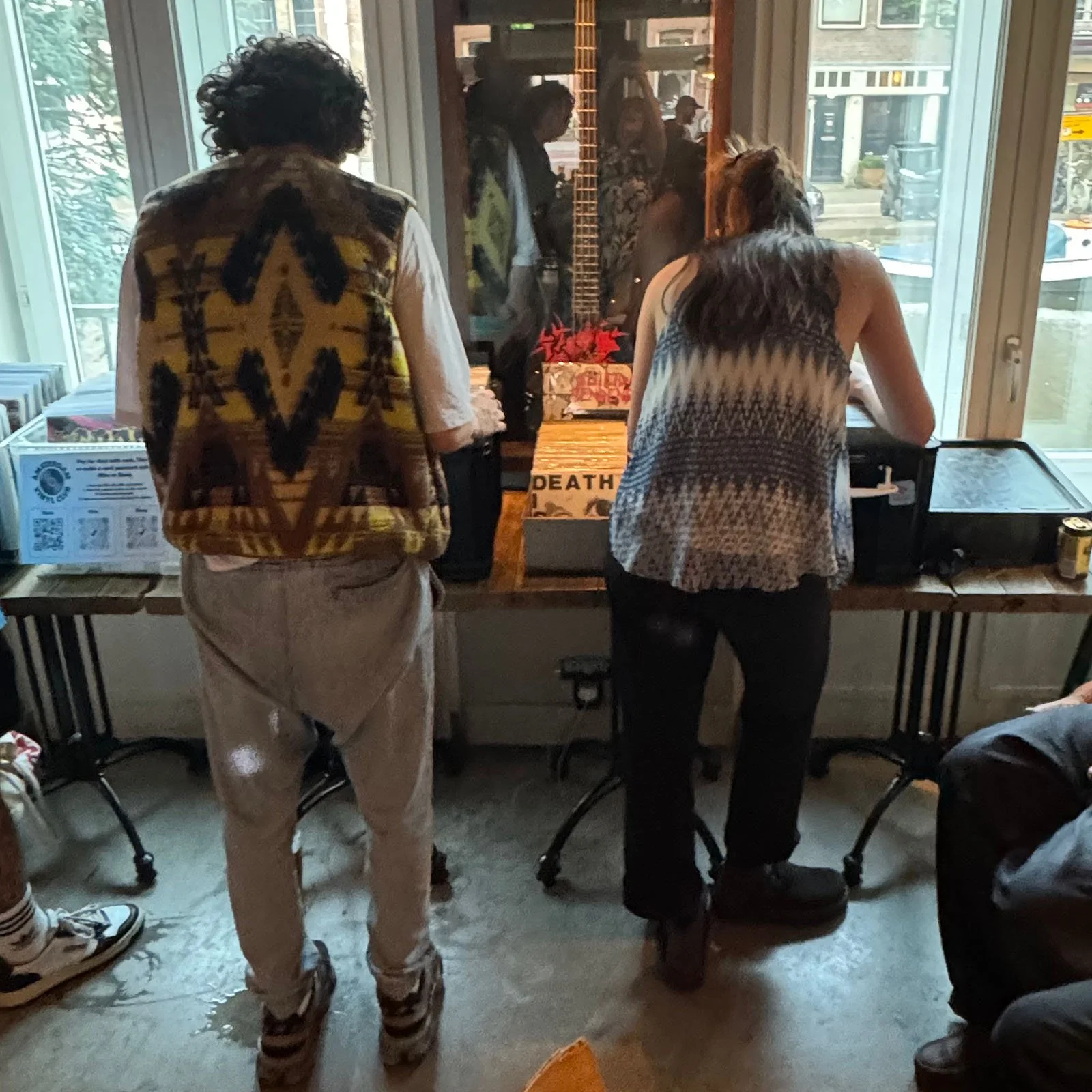 Two people browse records at an Amsterdam Vinyl Club live event. Two men are visible from behind, one with curly hair and a patterned vest, the other with straight hair and a sleeveless top.
