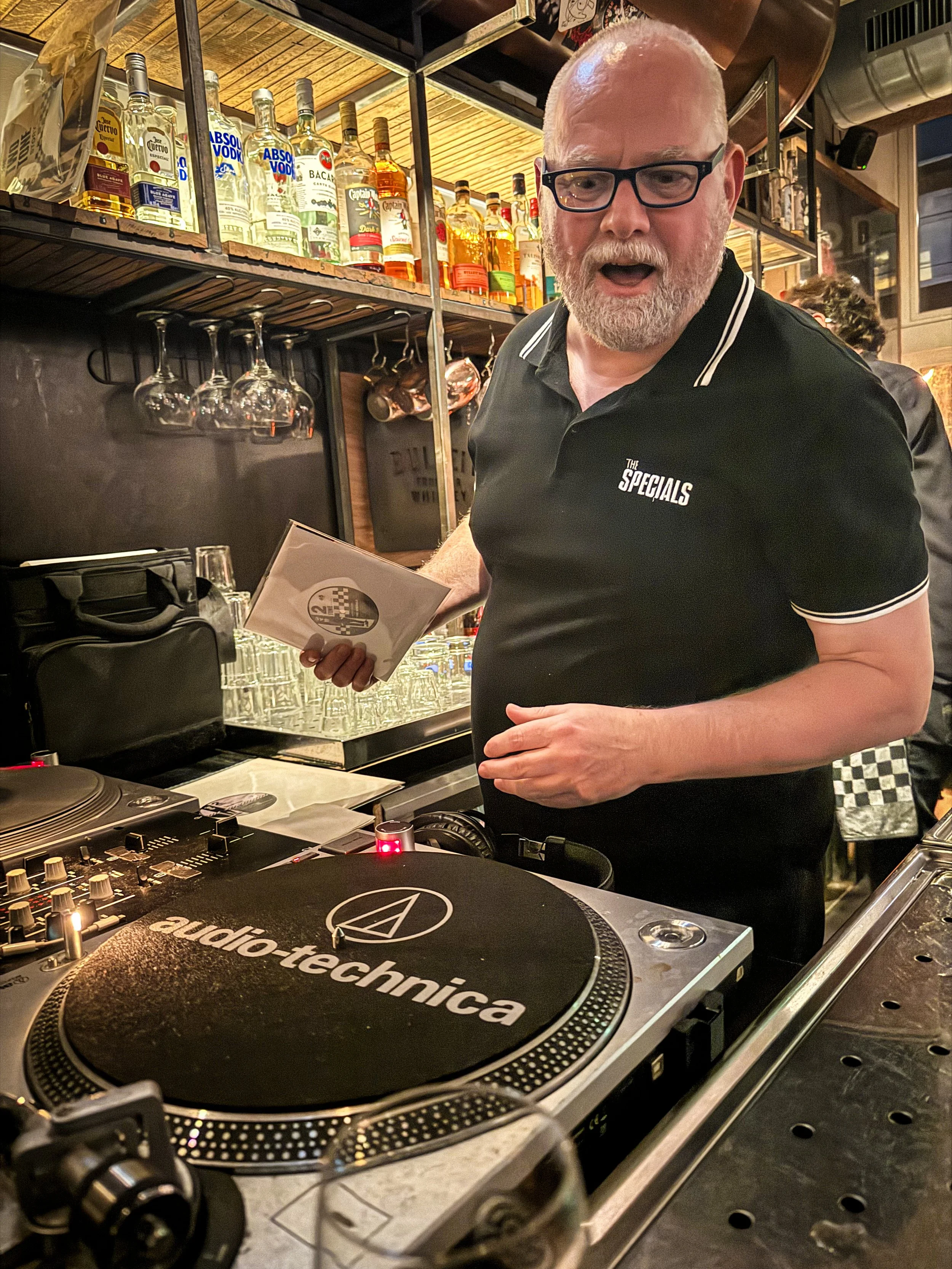 A man with glasses and a beard, wearing a black polo shirt with white trim, stands behind a DJ turntable, holding a small booklet. The background shows shelves with liquor bottles and hanging wine glasses in a bar or restaurant setting.